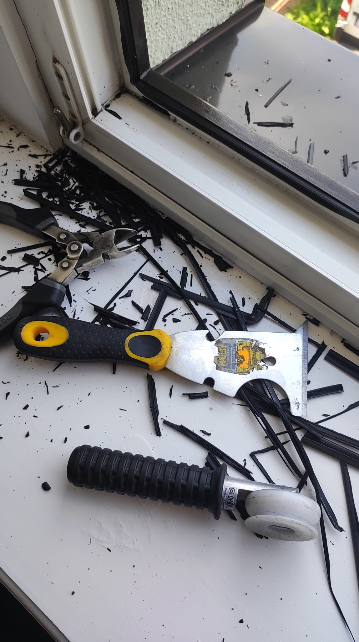 Demontaż uszczelki okiennej: białe okno, narzędzia (skrobak, szczypce, wałek dociskowy) i fragmenty czarnej uszczelki na białym parapecie. Praca w toku.