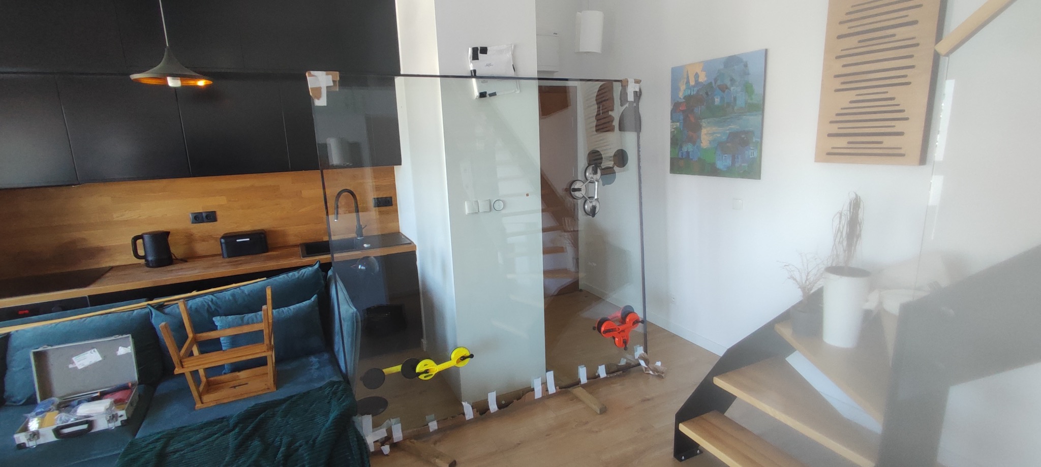 Montaż szklanej ścianki działowej w mieszkaniu, widoczne przyssawki i zabezpieczenia. W tle nowoczesna kuchnia i schody.