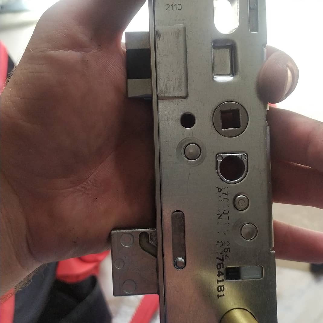 Detal okucia okiennego w dłoni, widoczne elementy zamka i mechanizmu. Metalowa powierzchnia z widocznymi oznaczeniami i numerami seryjnymi. Precyzyjne ujęcie.