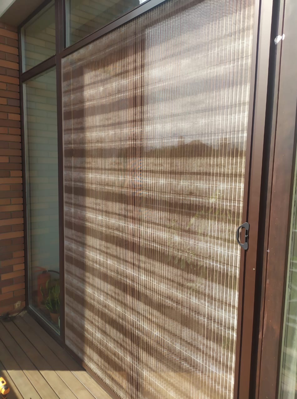 Zewnętrzna roleta plisowana w brązowej ramie, zamontowana na oknie budynku z cegły. Widoczny fragment drewnianego tarasu.