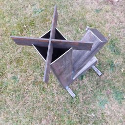 Serwis RN - Surowy, metalowy piec rakietowy z krzyżowym podajnikiem paliwa, spawany z profili kwadratowych, ustawiony na trawie. Widok z góry.