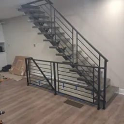 Nowoczesne schody metalowe z czarną balustradą w jasnym wnętrzu. Widoczne narzędzia i karton sugerują trwający montaż.