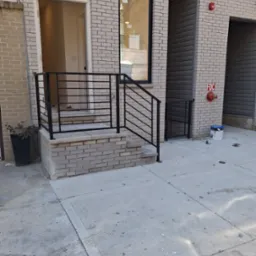 Metalowa balustrada schodowa w czarnym kolorze, zamontowana przed wejściem do budynku z cegły. Widoczny fragment chodnika i rośliny w donicy.