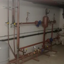 Kompleksowy system rur miedzianych z zaworami, manometrem i zbiornikiem wyrównawczym, zainstalowany w piwnicy. Widoczne mocowania do ściany i sufitu.