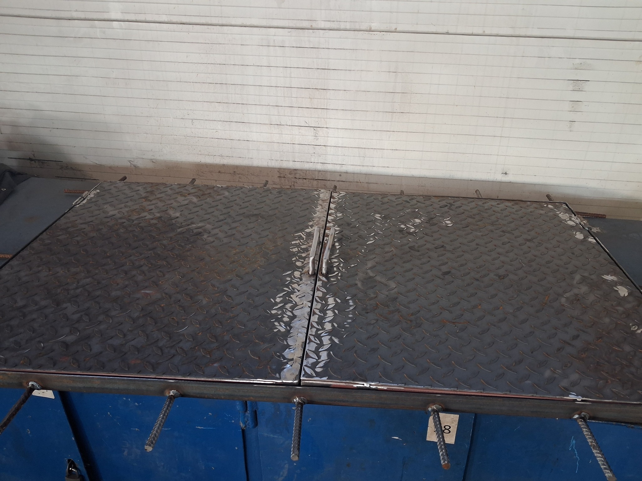 Dwu-częściowa, metalowa klapa z uchwytami, spawana z prętów, o powierzchni z blachy ryflowanej, prawdopodobnie właz lub pokrywa studni, na tle białej ściany.