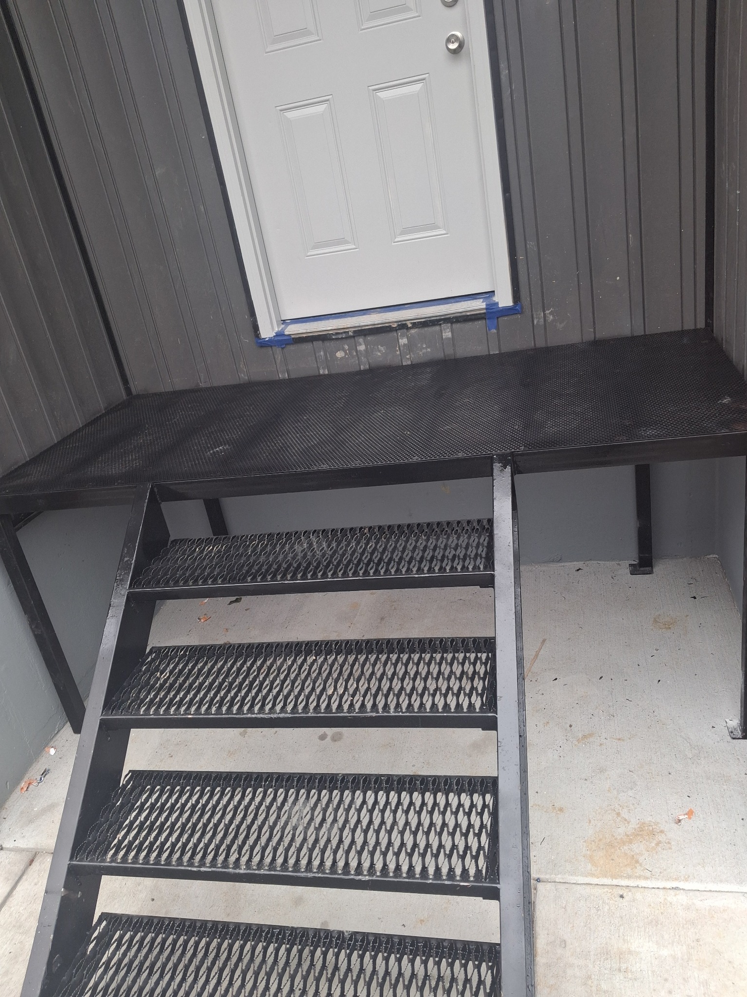 Metalowe schody zewnętrzne prowadzące do białych drzwi wejściowych, z siatkowymi stopniami, pomalowane na czarno, widok z dołu.