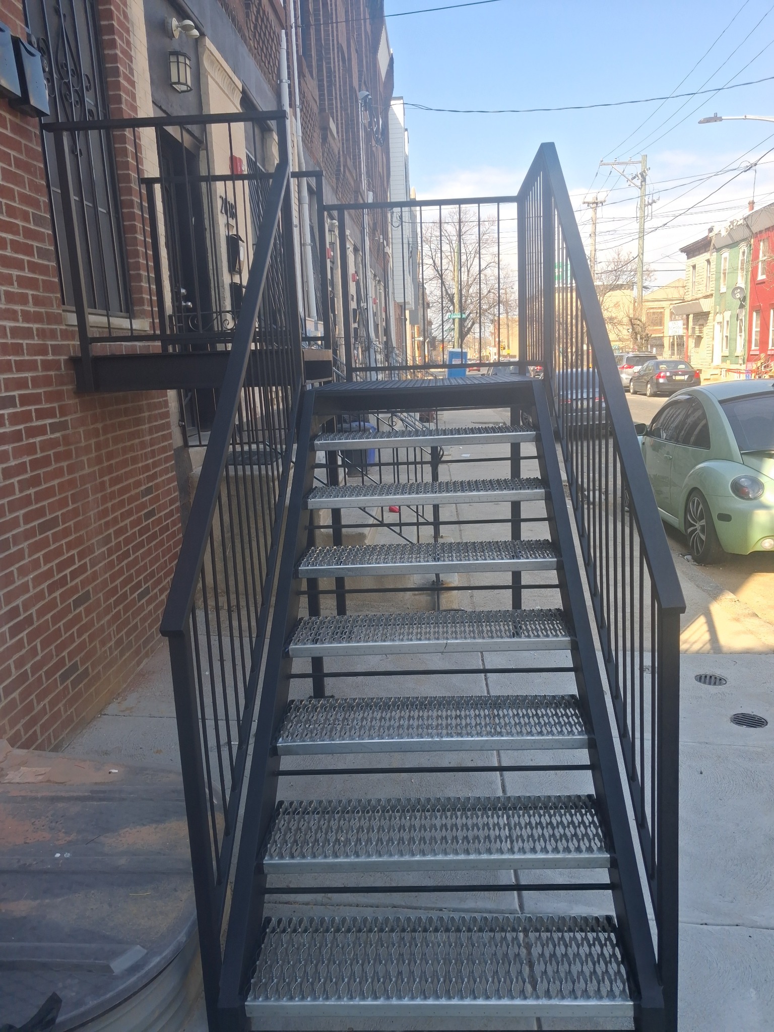 Zewnętrzne schody metalowe z balustradą, prowadzące do budynku z cegły. Schody mają ażurową konstrukcję, widoczne otoczenie miejskie i zaparkowany samochód.