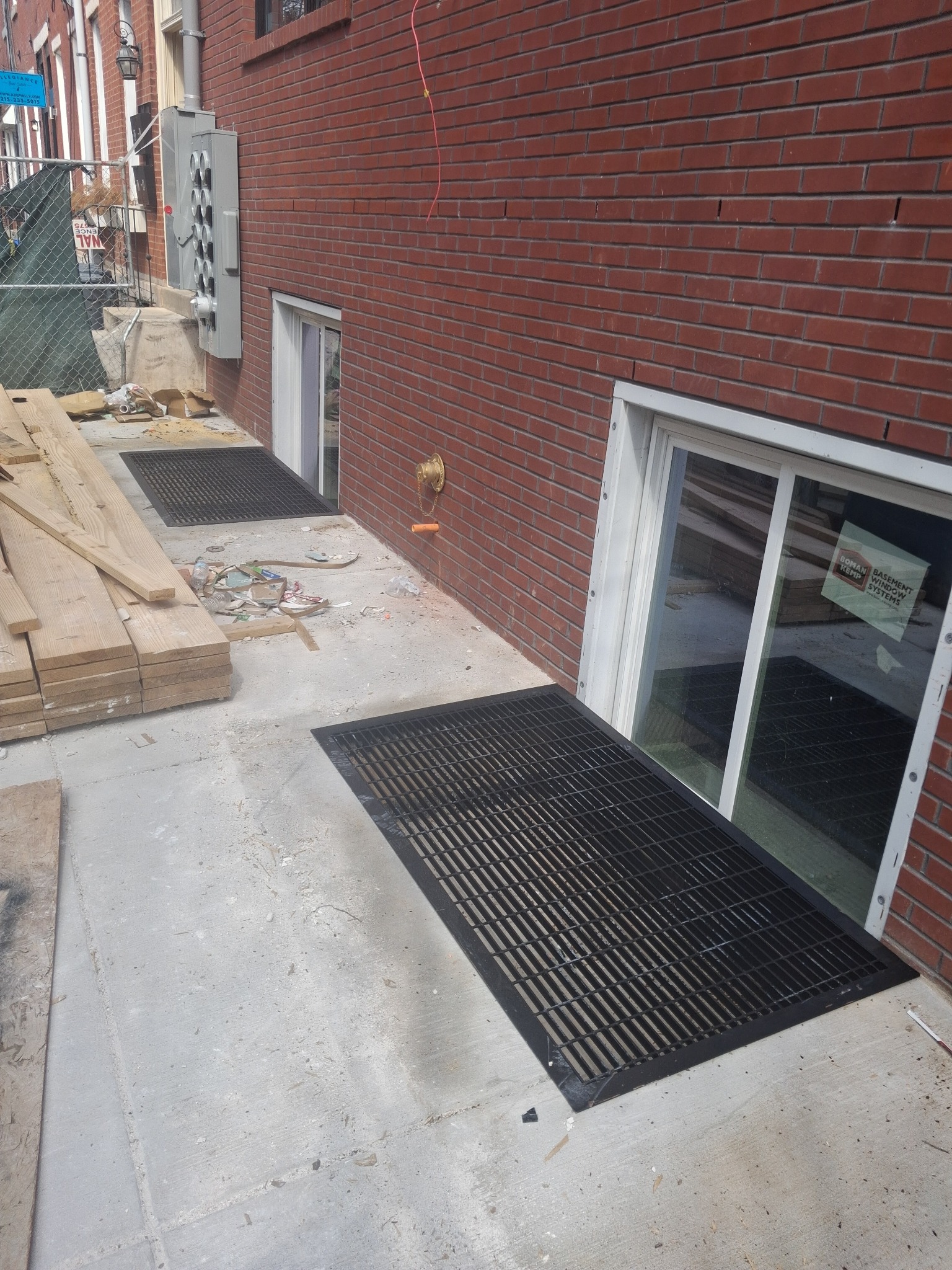 Montaż krat okiennych piwnicznych przy ścianie budynku z czerwonej cegły. Widoczne okna piwniczne i sterta desek. Kąt ujęcia z dołu w prawo.