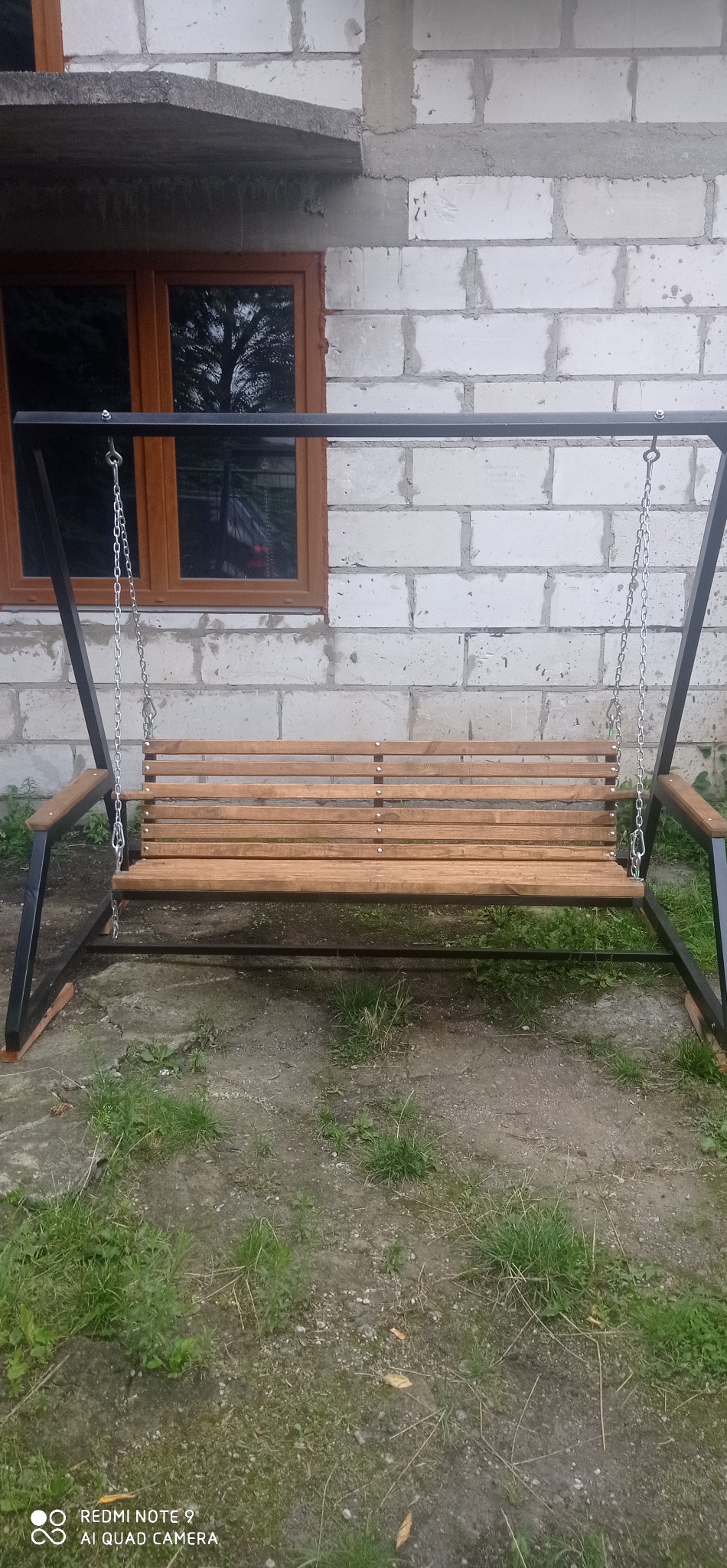 Metalowa huśtawka ogrodowa z drewnianym siedziskiem, zawieszona na łańcuchach, na tle ściany z białej cegły i okna z brązową ramą. Widok z przodu, na trawniku.