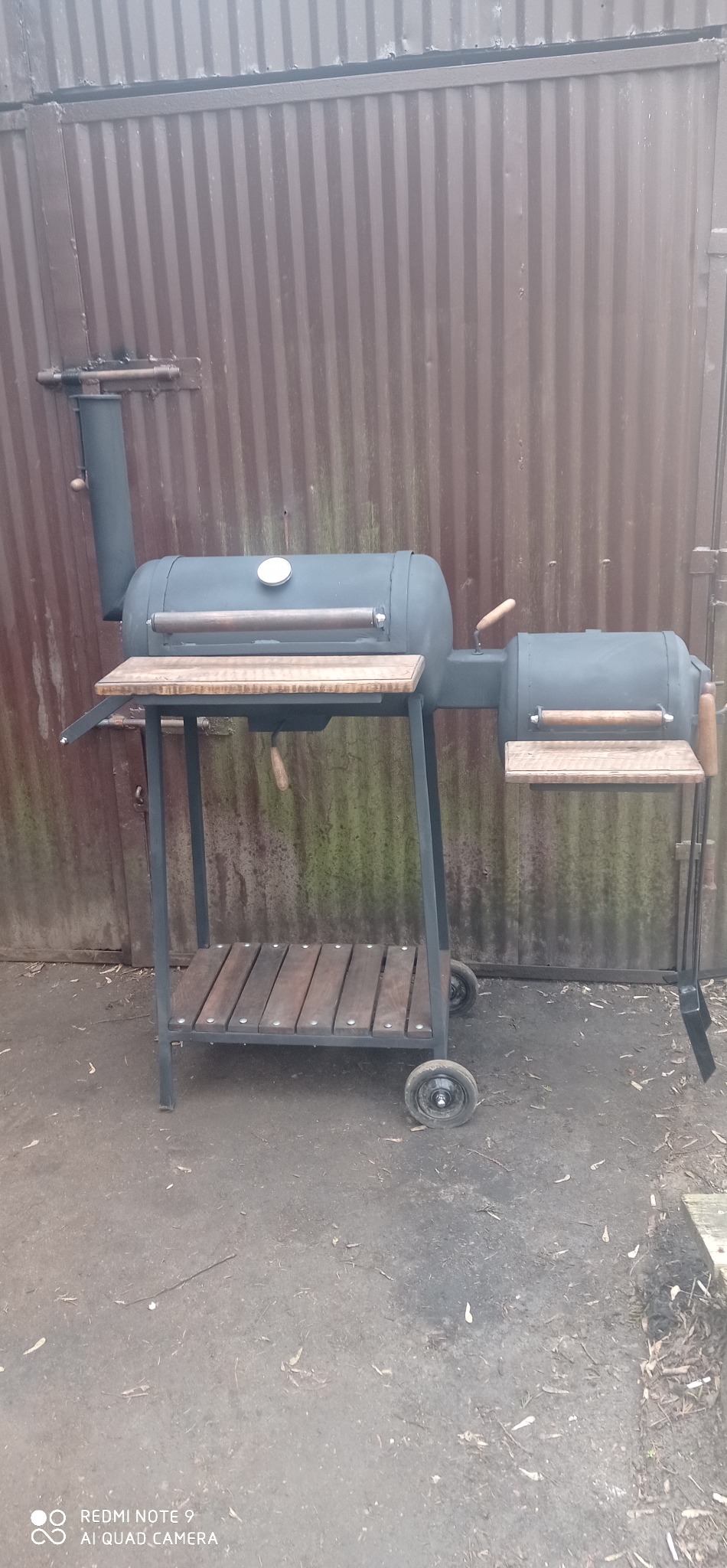 Czarny grill z metalu i drewna, na kółkach, z drewnianymi blatami roboczymi, na tle blaszanej ściany. Widoczne detale konstrukcyjne.