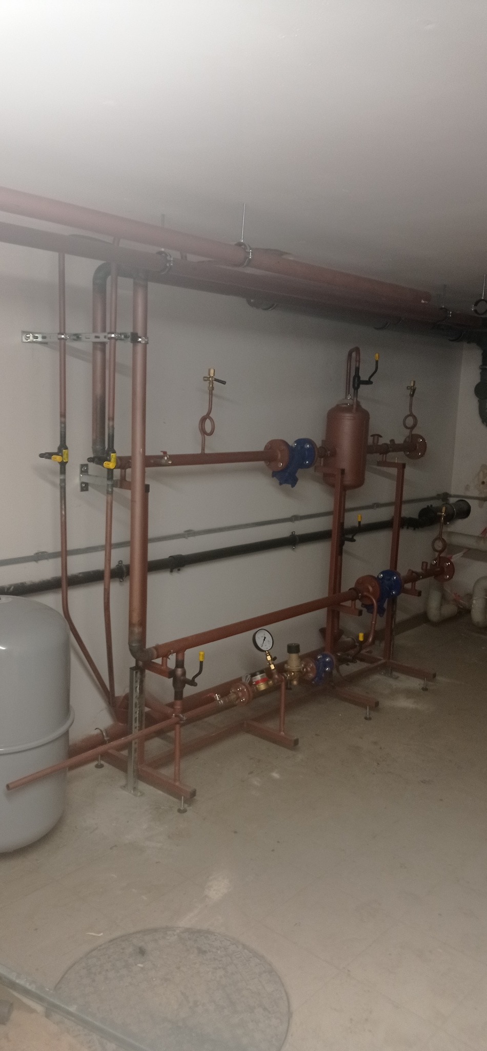 Kompleksowy system rur miedzianych z zaworami, manometrem i zbiornikiem wyrównawczym, zainstalowany w piwnicy. Widoczne mocowania do ściany i sufitu.