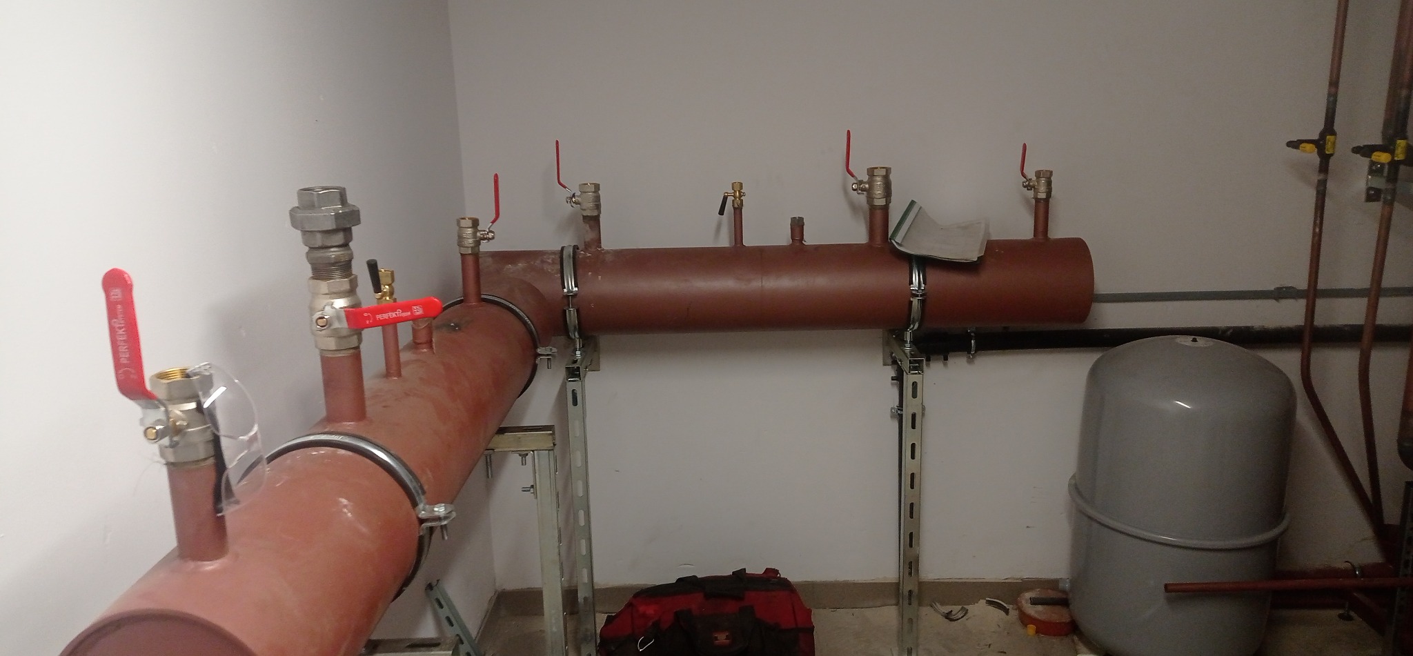 Brązowe rury instalacji CO z zaworami, zamontowane na metalowych wspornikach przy białej ścianie. Widoczny szary zbiornik wyrównawczy i torba narzędziowa na podłodze.