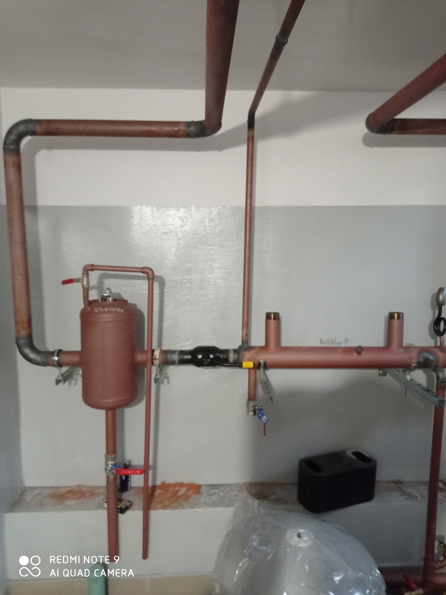 Miedziana instalacja grzewcza z zaworami i zbiornikiem wyrównawczym na tle szarej ściany. Widoczne spawane łączenia rur.