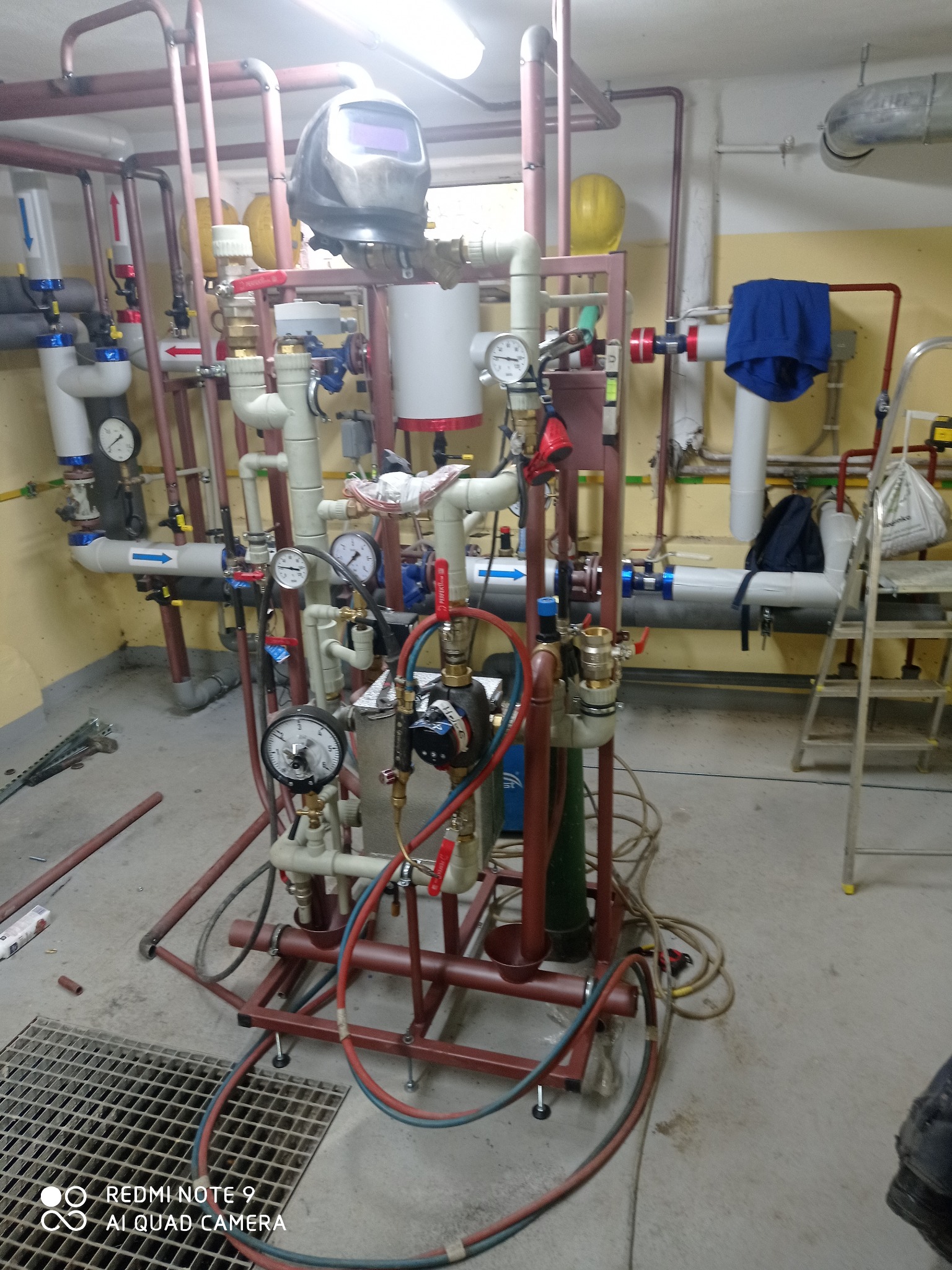 Skomplikowana instalacja hydrauliczna z licznymi rurami, manometrami i zaworami, widoczna maska spawalnicza i butla z gazem, sugerująca etap prac instalacyjnych.