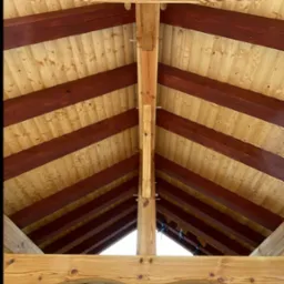 Wnętrze drewnianej altany z widoczną konstrukcją dachu, bejcowanymi belkami i naturalnym rysunkiem drewna. W tle zieleń i budynek.