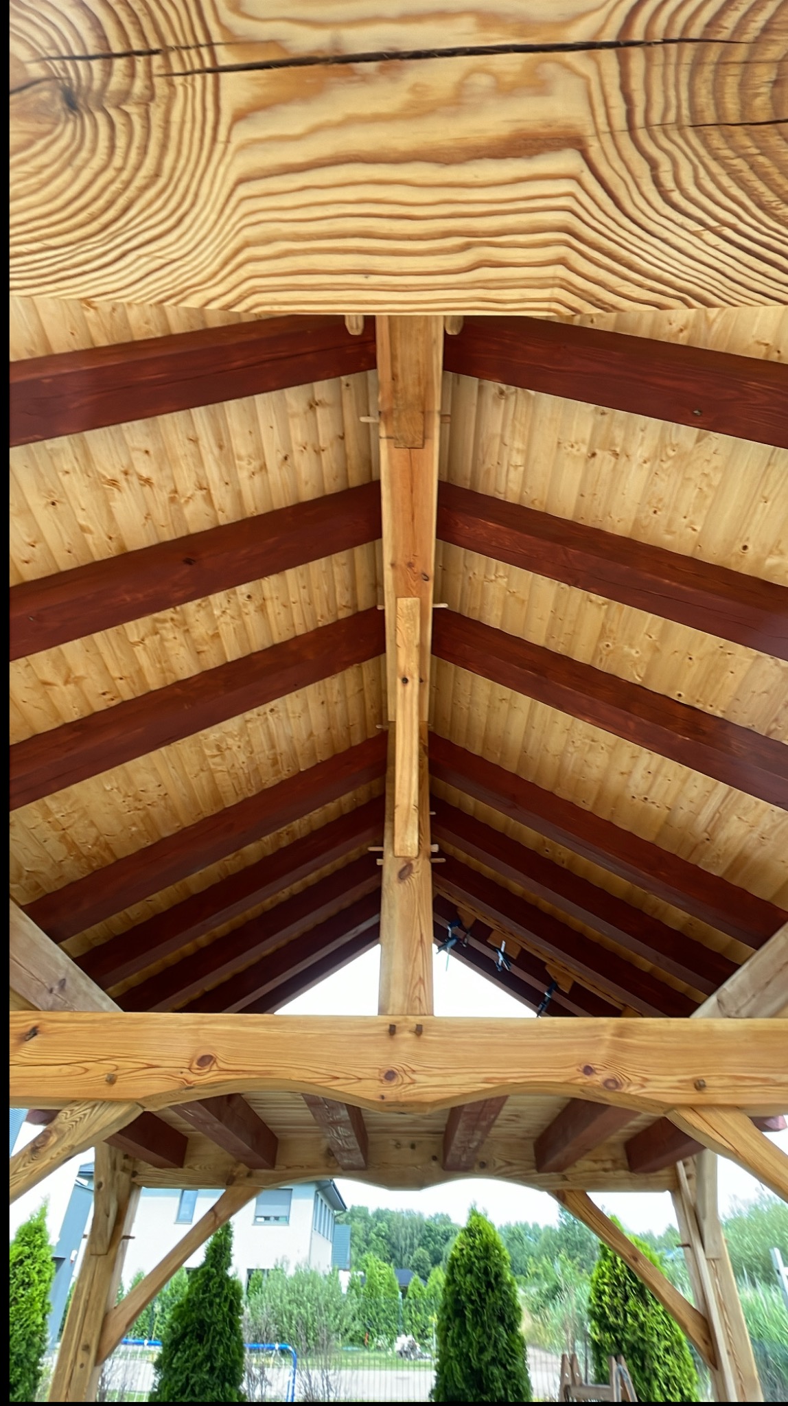 Wnętrze drewnianej altany z widoczną konstrukcją dachu, bejcowanymi belkami i naturalnym rysunkiem drewna. W tle zieleń i budynek.