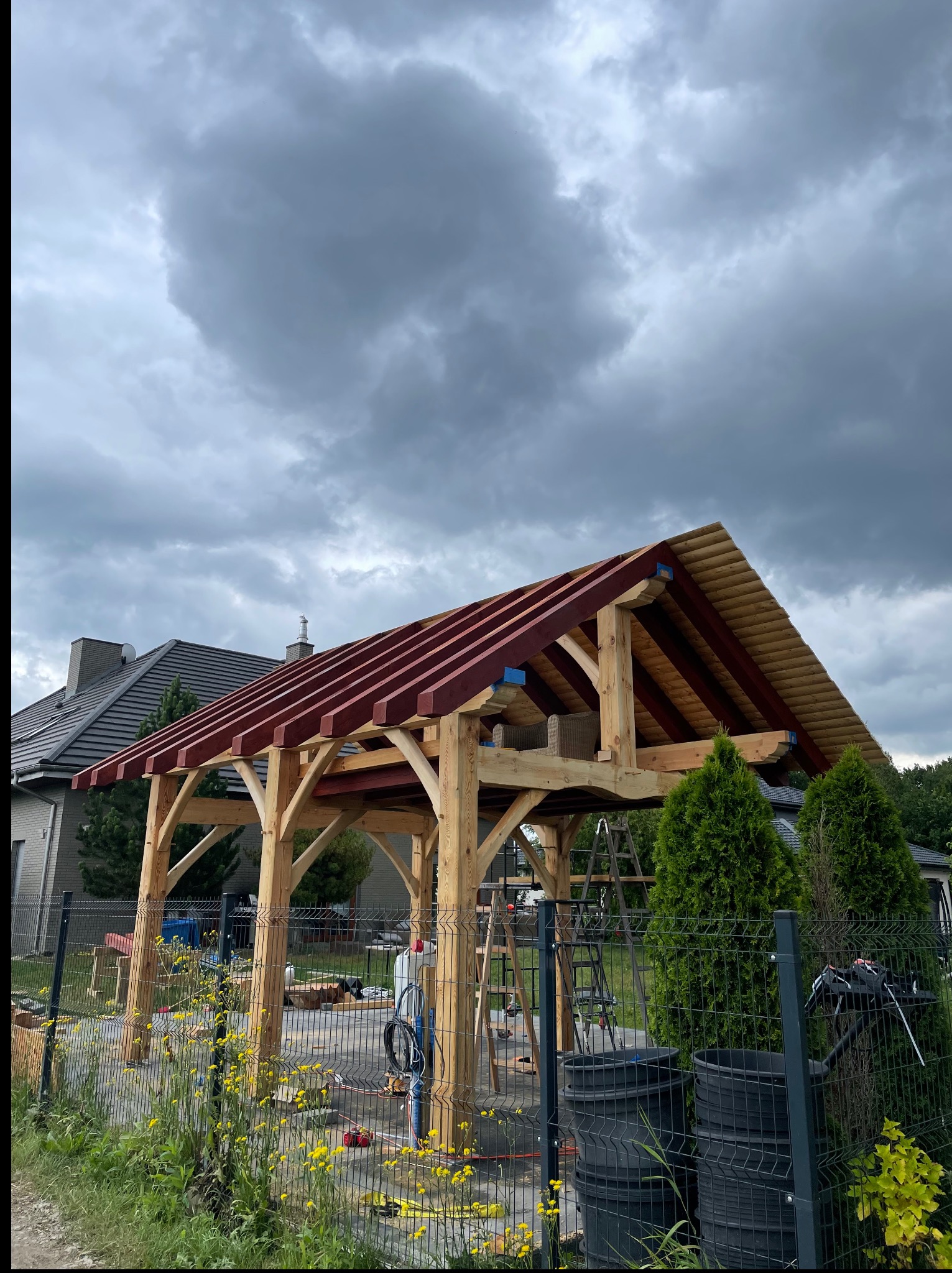 Wznoszenie drewnianej konstrukcji altany z czerwonymi belkami na tle pochmurnego nieba. Widoczne narzędzia i drabina, sugerujące etap budowy.