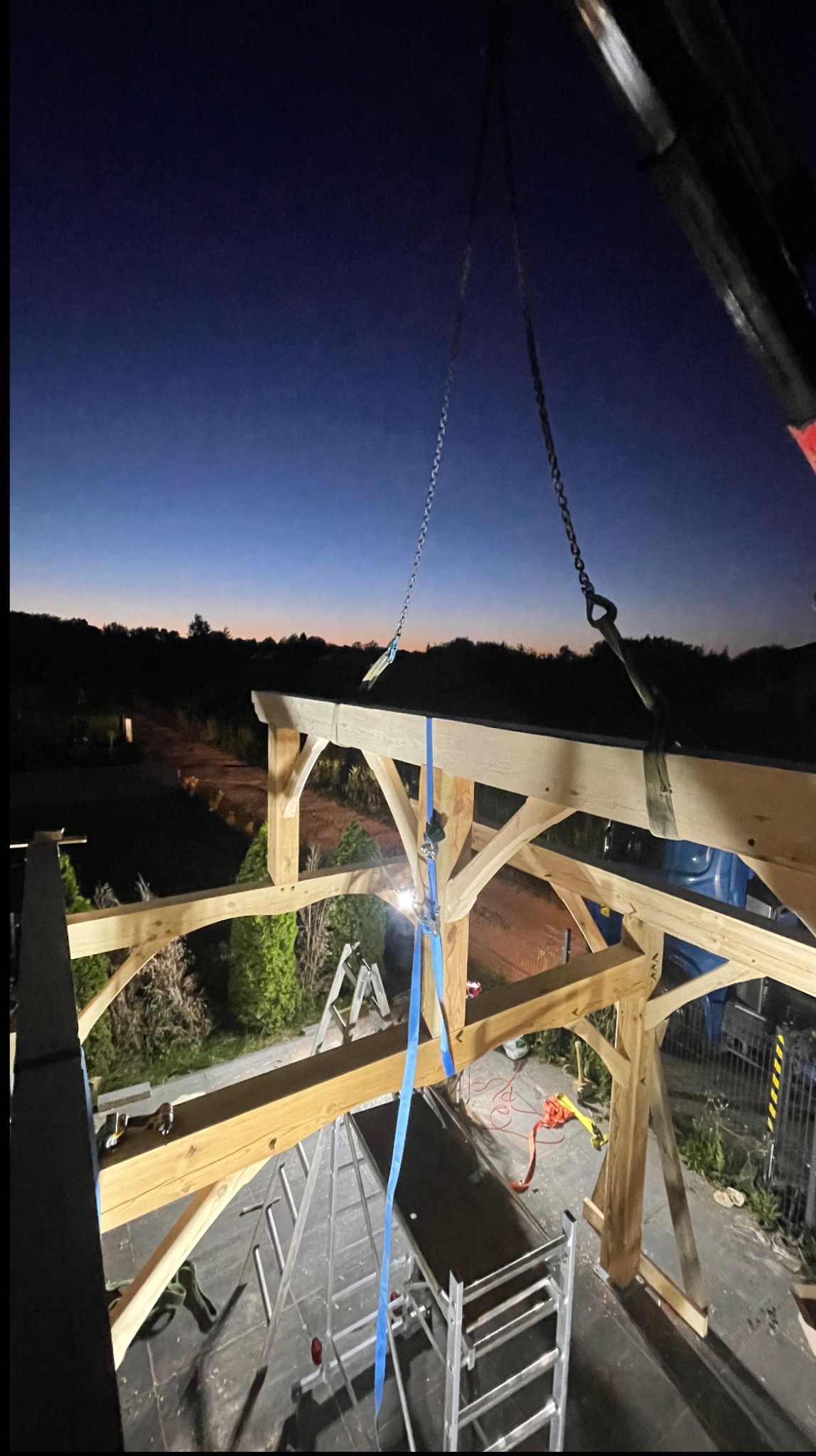Montaż drewnianej konstrukcji: widok z góry na szkielet z belek, podnośnik i drabiny na tle wieczornego nieba.