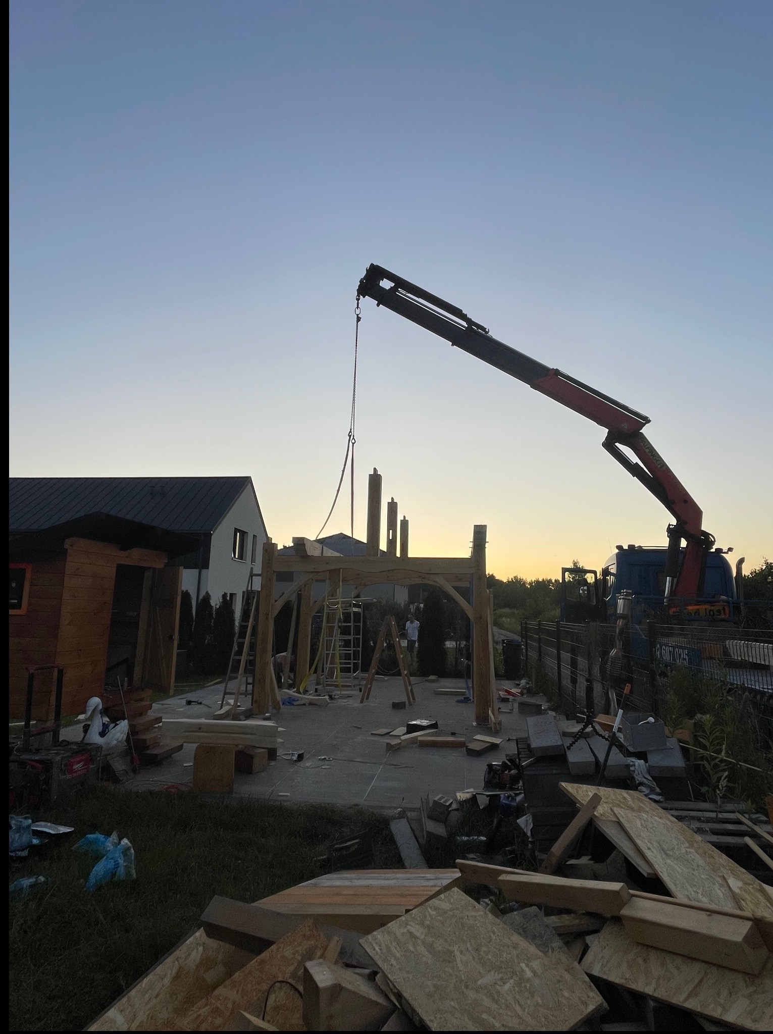 Montaż drewnianej konstrukcji z użyciem dźwigu na tle zachodzącego słońca. Elementy drewniane, plac budowy i ciężarówka widoczne.