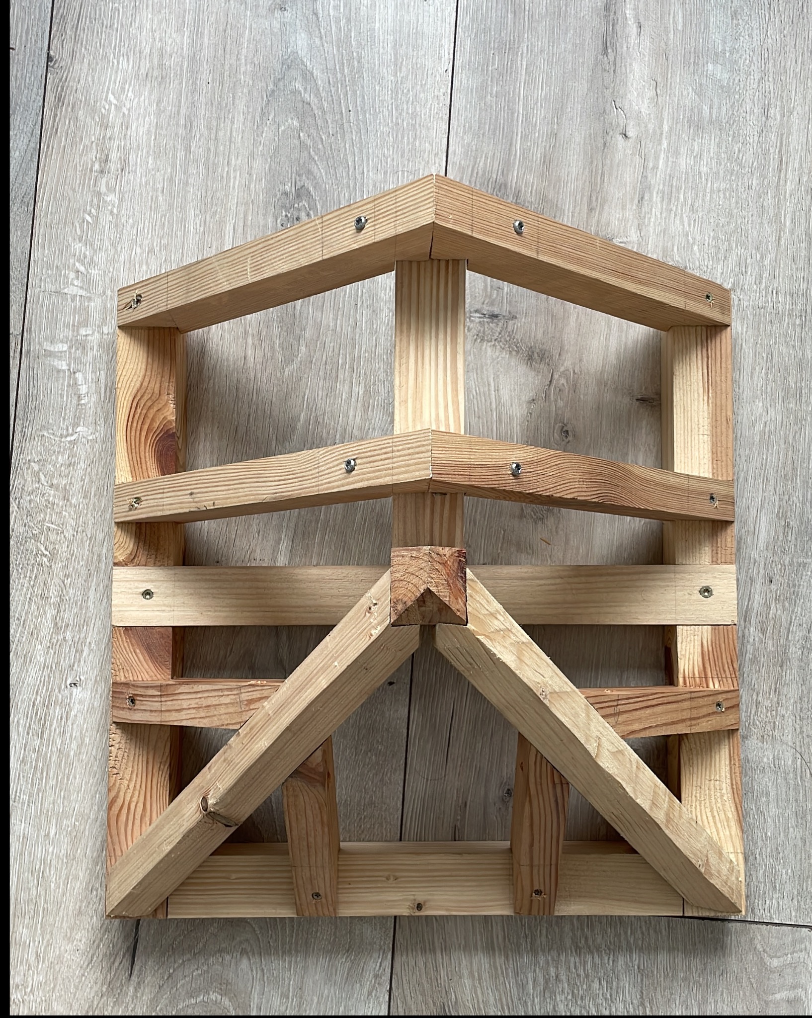 Surowe drewniane elementy konstrukcyjne, złączone śrubami, tworzące ramę przypominającą fragment altany lub pergoli, na tle drewnopodobnej powierzchni.