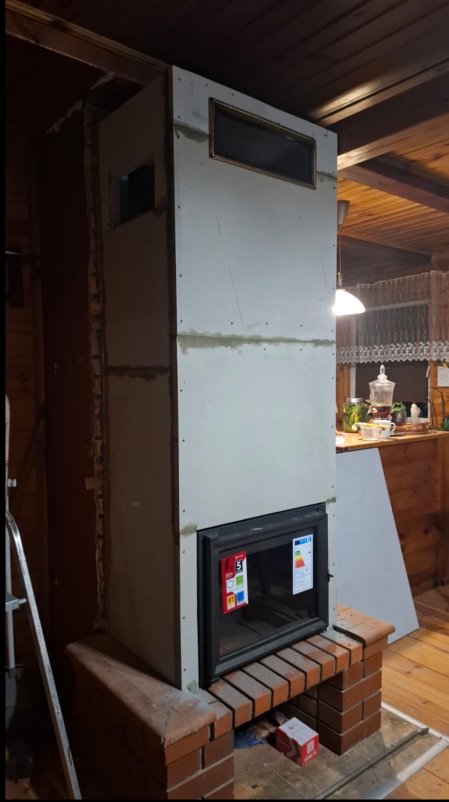 Wnętrze z kominkiem w trakcie obudowy płytami gipsowymi, widoczne ceglane elementy i drabina, sugerujące etap prac wykończeniowych.