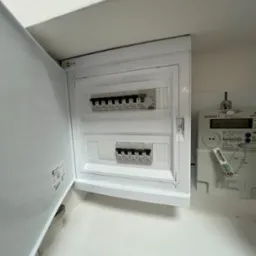 Otwarta, biała skrzynka z bezpiecznikami marki Legrand, zamontowana na ścianie obok licznika energii elektrycznej Norax 1.