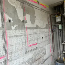 Ściana w trakcie remontu z wyżłobieniami pod kable, zaznaczonym przebiegiem instalacji r&oacute;żowym sprayem i otworami pod puszki elektryczne. Widoczne rury i instalacja gazowa.