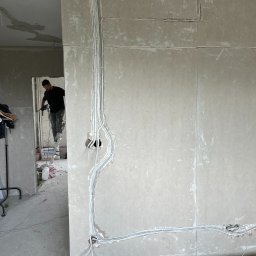 PT4Y GROUP SPÓŁKA Z OGRANICZONĄ ODPOWIEDZIALNOŚCIĄ - Ściana z instalacją elektryczną w peszlach, monter na drabinie w tle, wiertarka w dłoni. Prace wykończeniowe w surowym wnętrzu. Widoczne przewody i puszki.