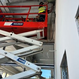 PT4Y GROUP SPÓŁKA Z OGRANICZONĄ ODPOWIEDZIALNOŚCIĄ - Elektryk w kamizelce na podnośniku montuje kable w metalowej korycie pod sufitem. Widoczne przewody i elementy instalacji. Praca na wysokości.