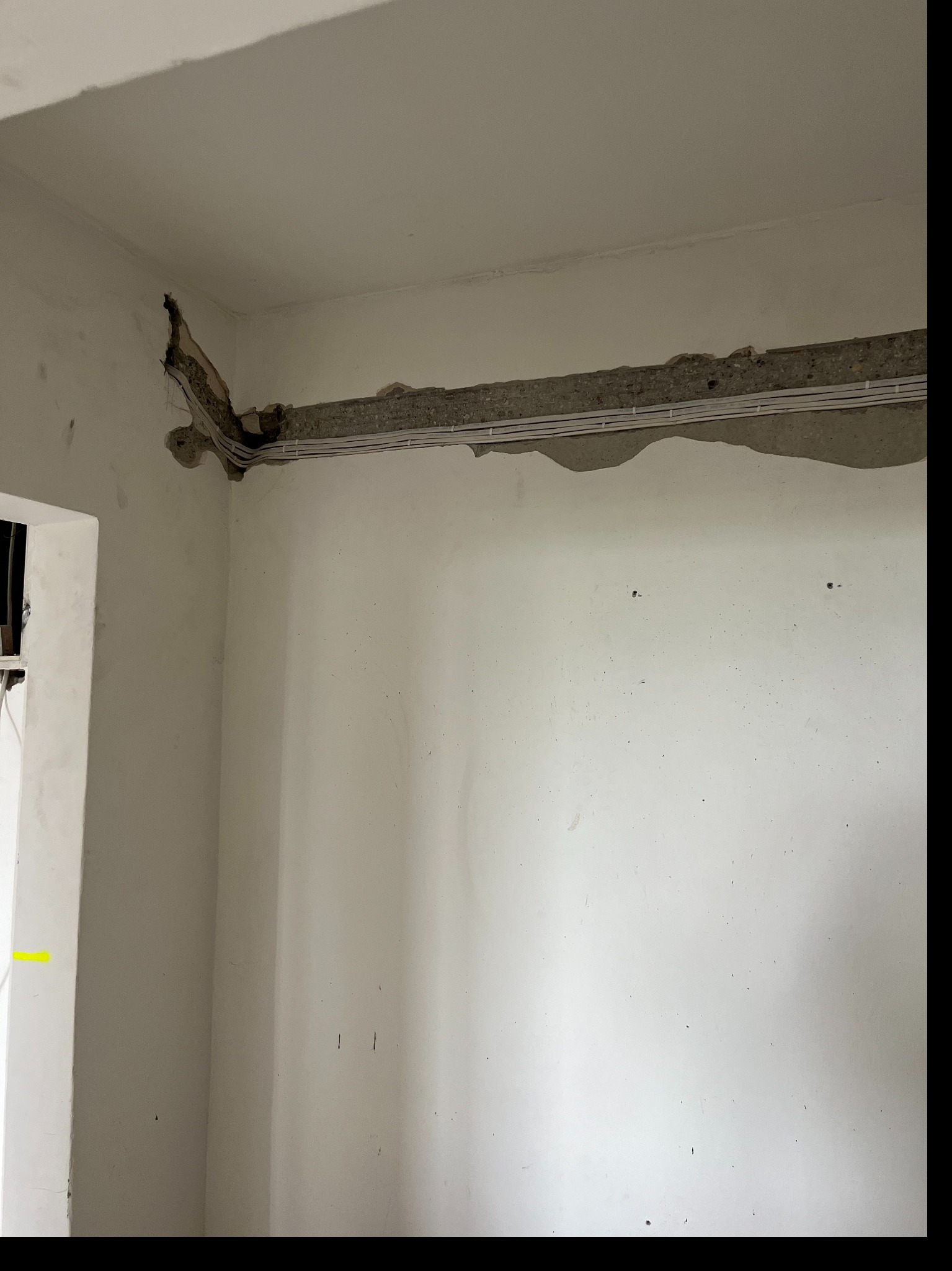 Białe przewody elektryczne ułożone w wyżłobieniu ściany, widoczne fragmenty betonu i instalacji w rogu pomieszczenia. Etap prac instalacyjnych.