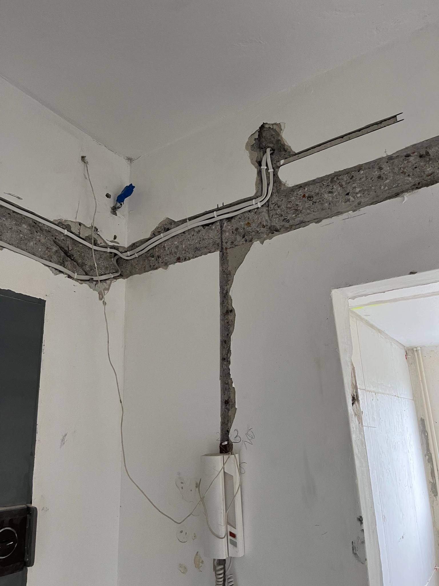 Białe kable elektryczne prowadzone w wyżłobieniach betonowej ściany w rogu pomieszczenia, widoczny domofon i fragment ramy drzwi.