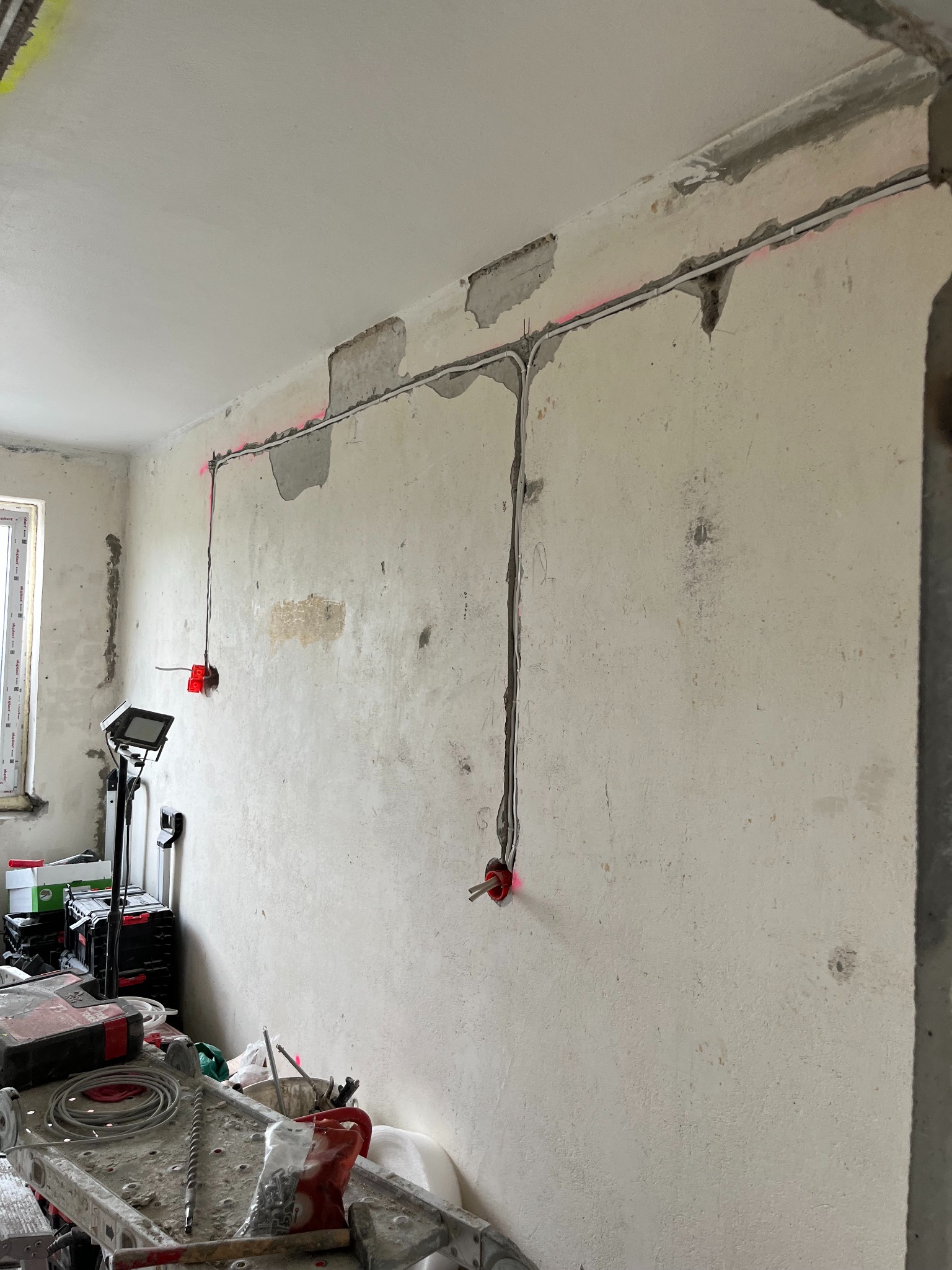 Montaż instalacji elektrycznej w surowym wnętrzu. Kable poprowadzone w bruzdach, czerwone puszki pod gniazdka, narzędzia pracy na drabinie. Widoczne ślady po laserze.