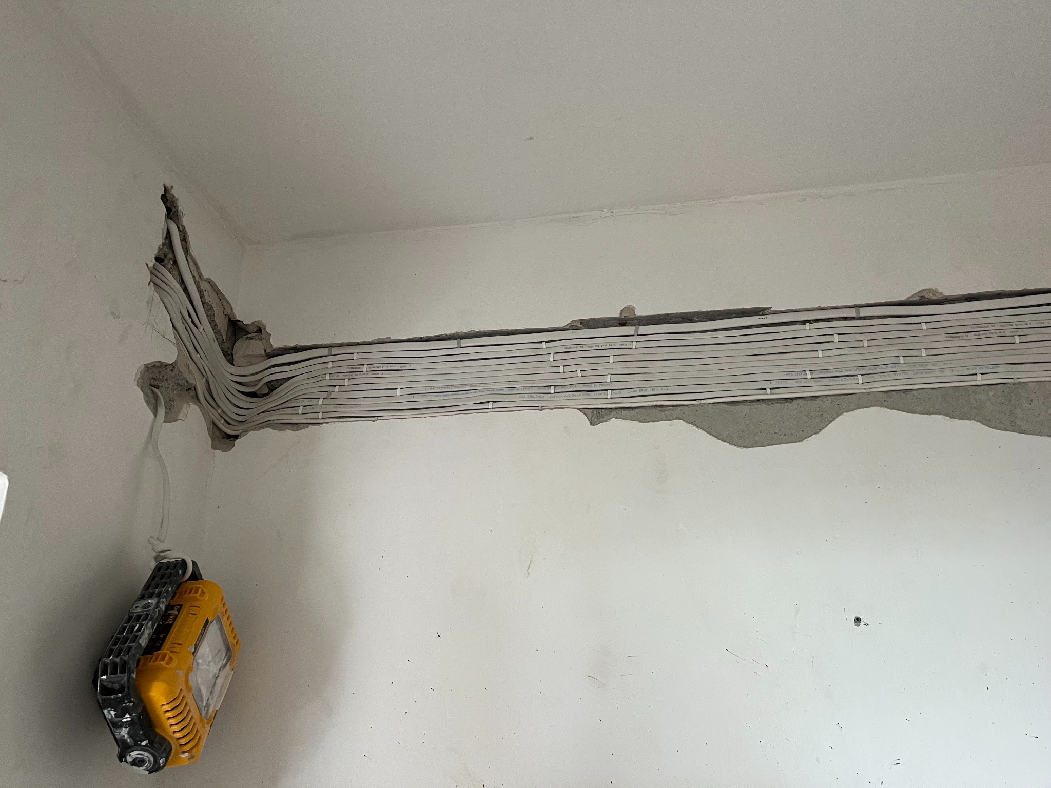 Białe przewody elektryczne ułożone w bruździe w ścianie, zabezpieczone klamrami. W rogu ściany widoczne miejsce ich wejścia. Na dole wisi lampa robocza.