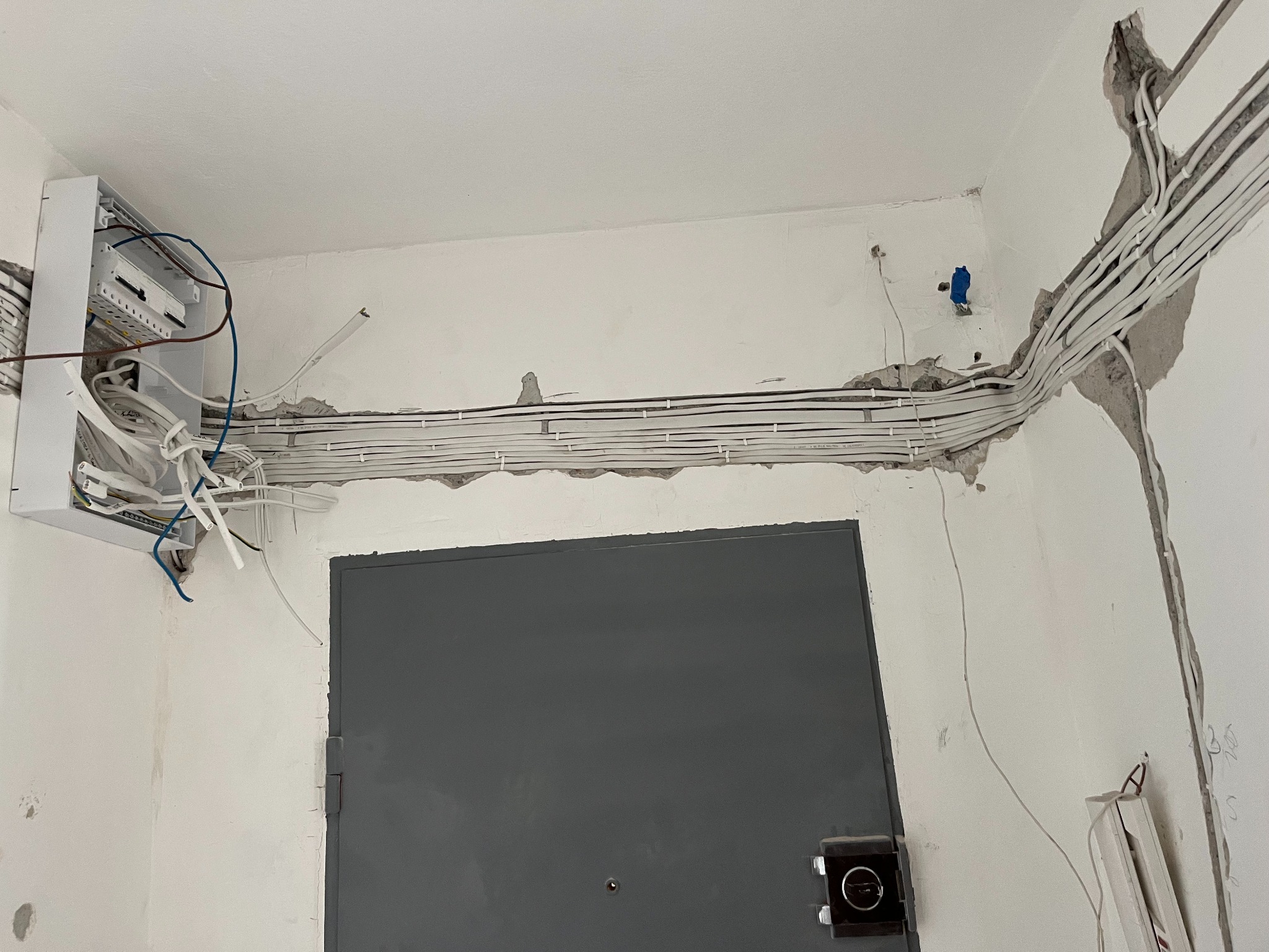 Rozprowadzenie przewodów elektrycznych w korytarzu, widoczna skrzynka rozdzielcza i przewody w bruzdach ściennych przed tynkowaniem. Szara metalowa futryna drzwi.