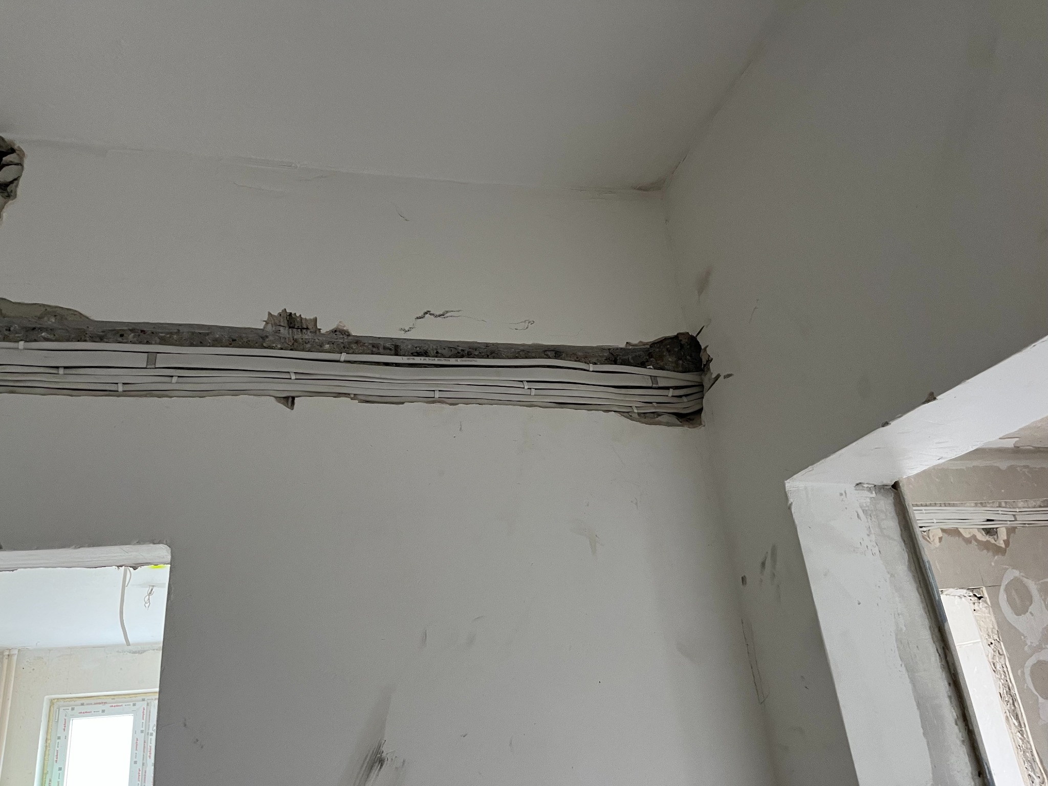 Białe przewody elektryczne ułożone w bruździe w ścianie, przygotowane do zakrycia tynkiem. Widoczne fragmenty ścian i sufitu w trakcie remontu.