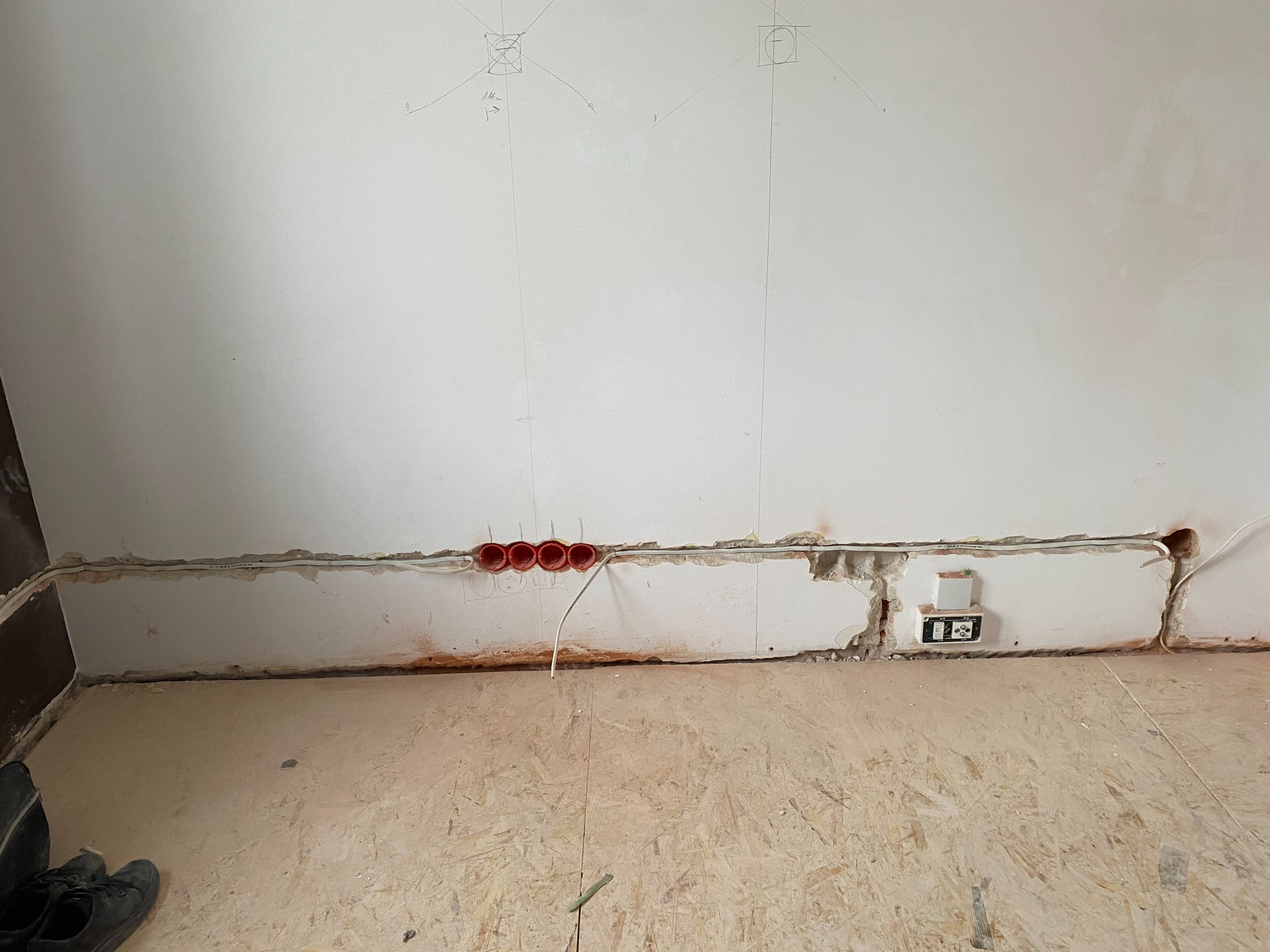 Wnęka w ścianie z przewodami i puszkami elektrycznymi, widoczne oznaczenia na ścianie. W tle surowe wykończenie wnętrza i podłoga z płyt OSB.