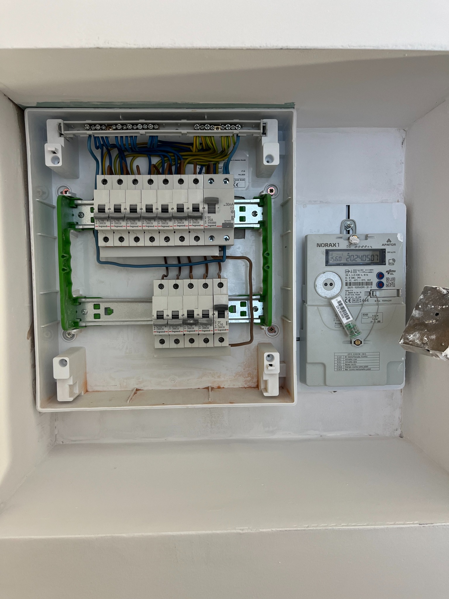 Rozdzielnia elektryczna z zamontowanym licznikiem energii, widoczne bezpieczniki i okablowanie. Świeżo pomalowana ściana z widocznymi śladami szpachli.