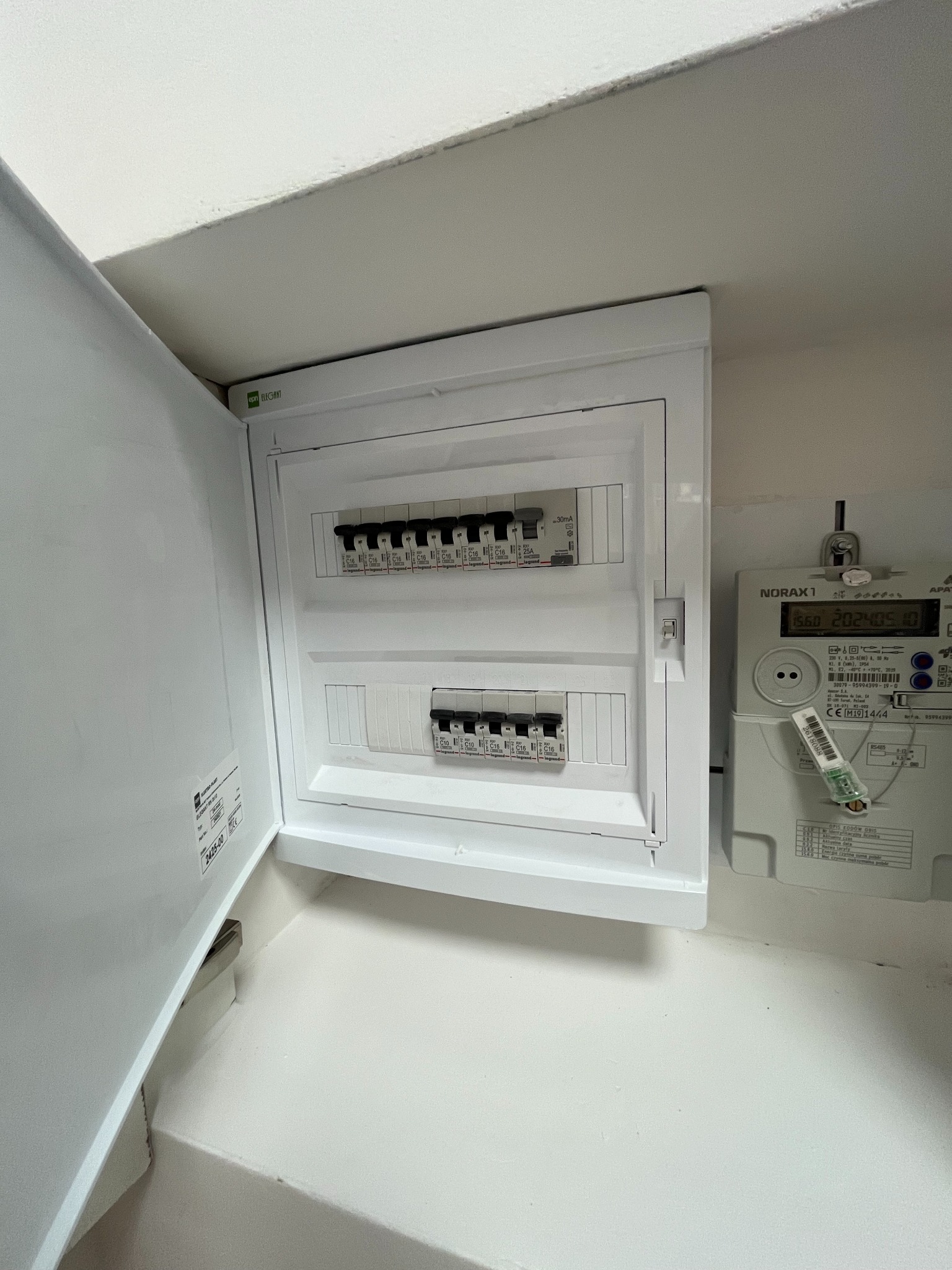 Otwarta, biała skrzynka z bezpiecznikami marki Legrand, zamontowana na ścianie obok licznika energii elektrycznej Norax 1.