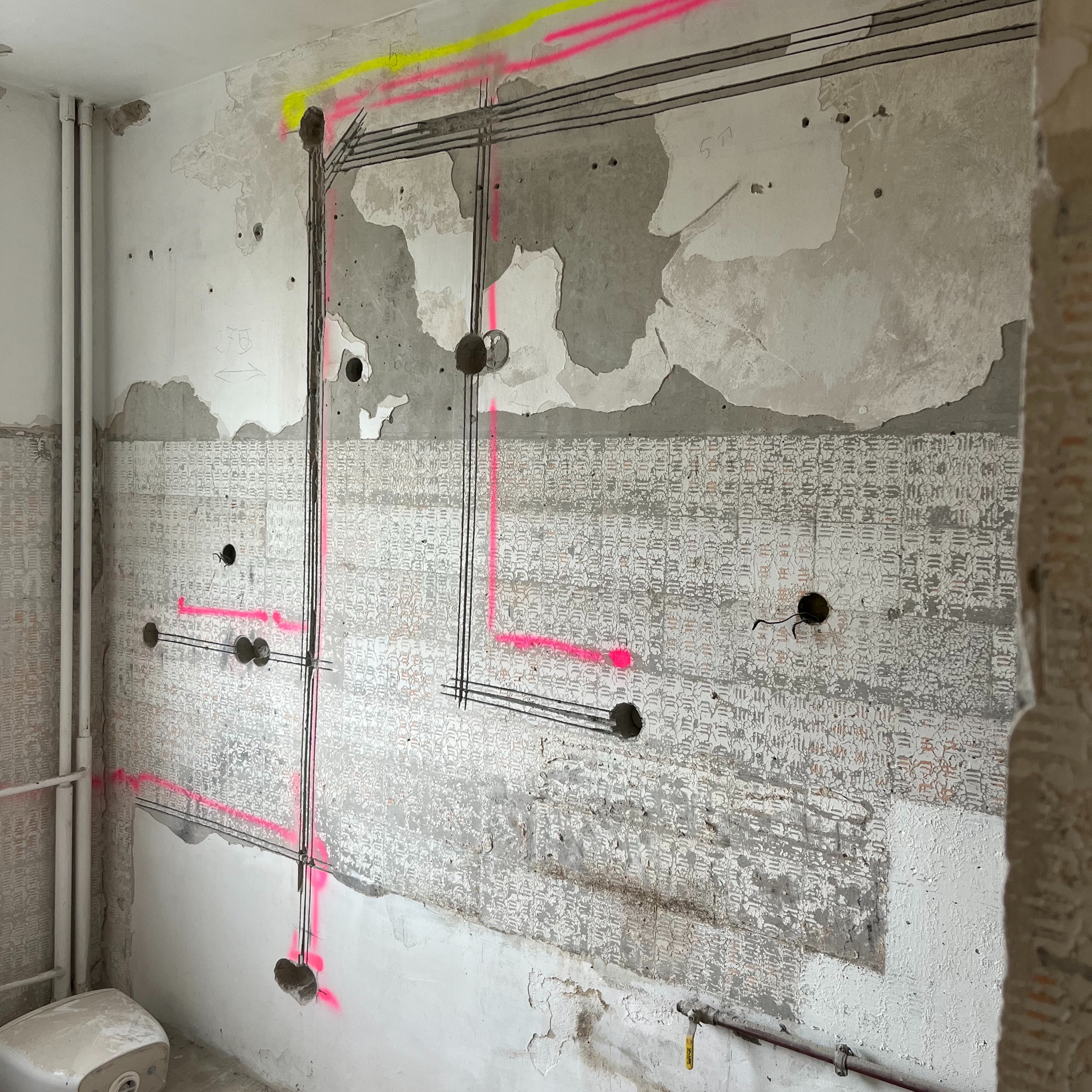 Ściana z wyznaczonymi trasami kabli elektrycznych, oznaczonymi różowym i żółtym sprayem, widoczne puszki instalacyjne i fragment instalacji wodnej.