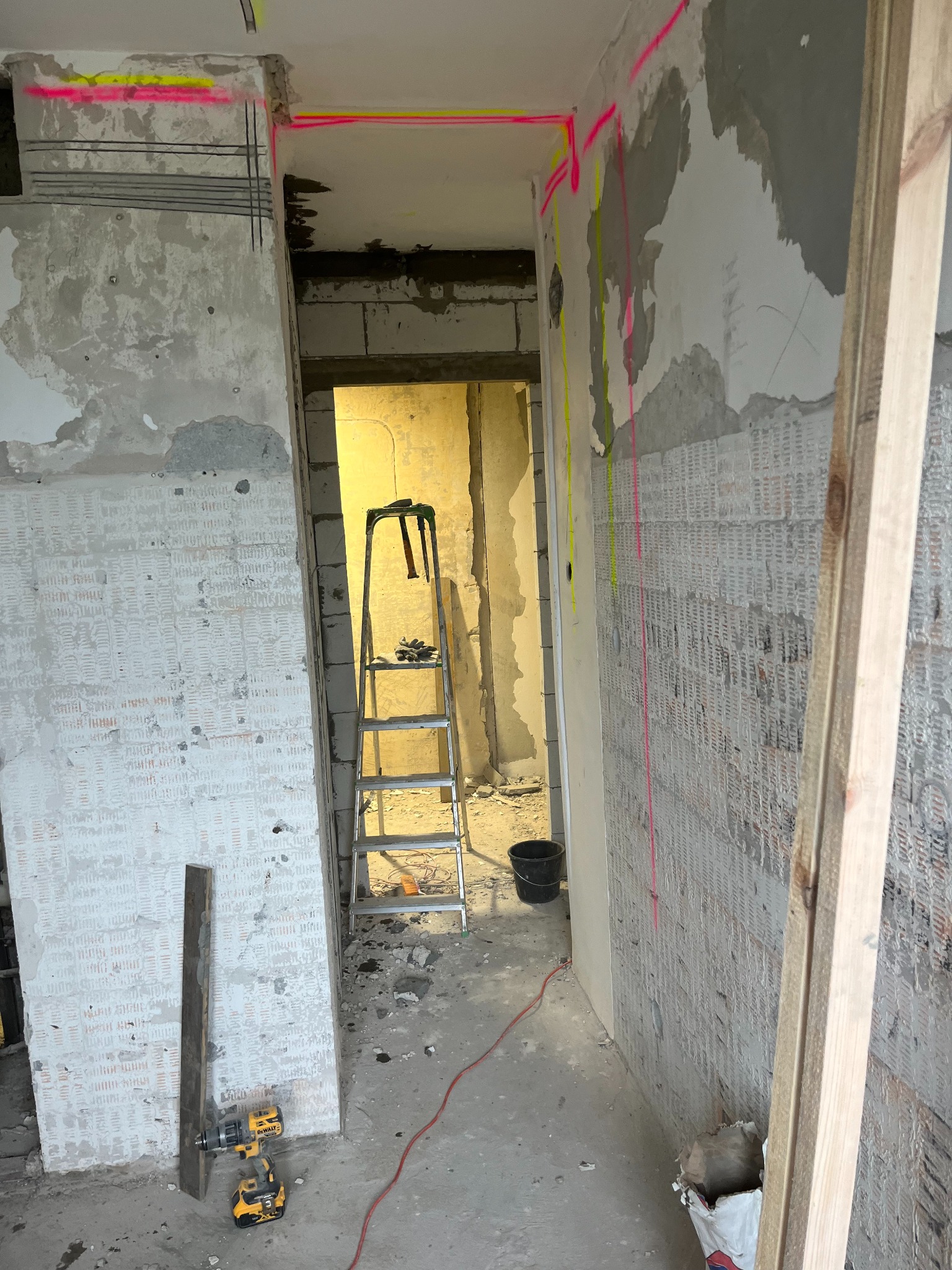 Wnętrze w trakcie remontu z widocznymi przewodami oznaczonymi neonowym kolorem, drabina i narzędzia. Ściany przygotowane do położenia instalacji.