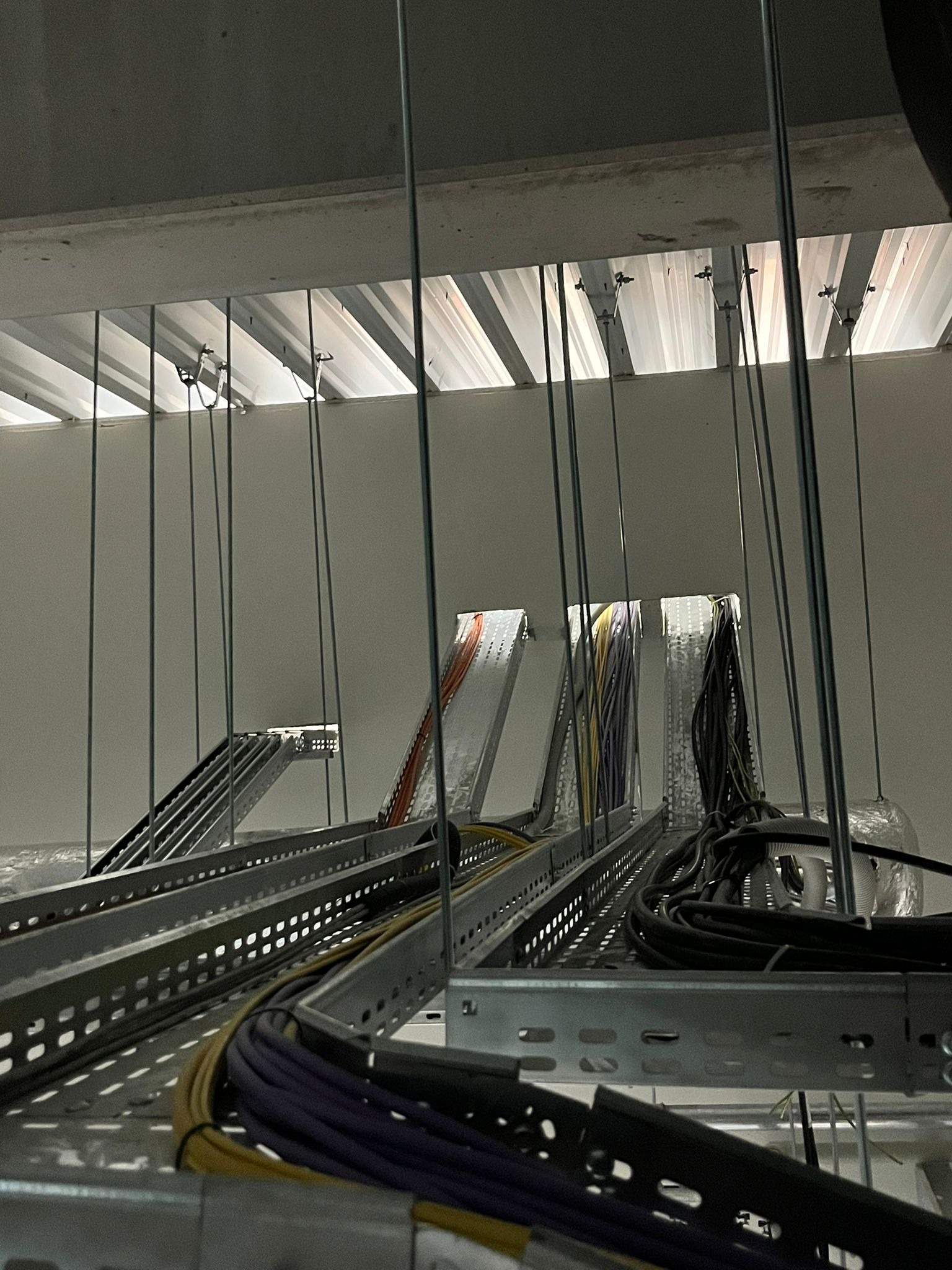 Koryta kablowe ze stalowymi wspornikami pod sufitem, prowadzące różnokolorowe przewody elektryczne. Instalacja w nowym budynku, widok z dołu.
