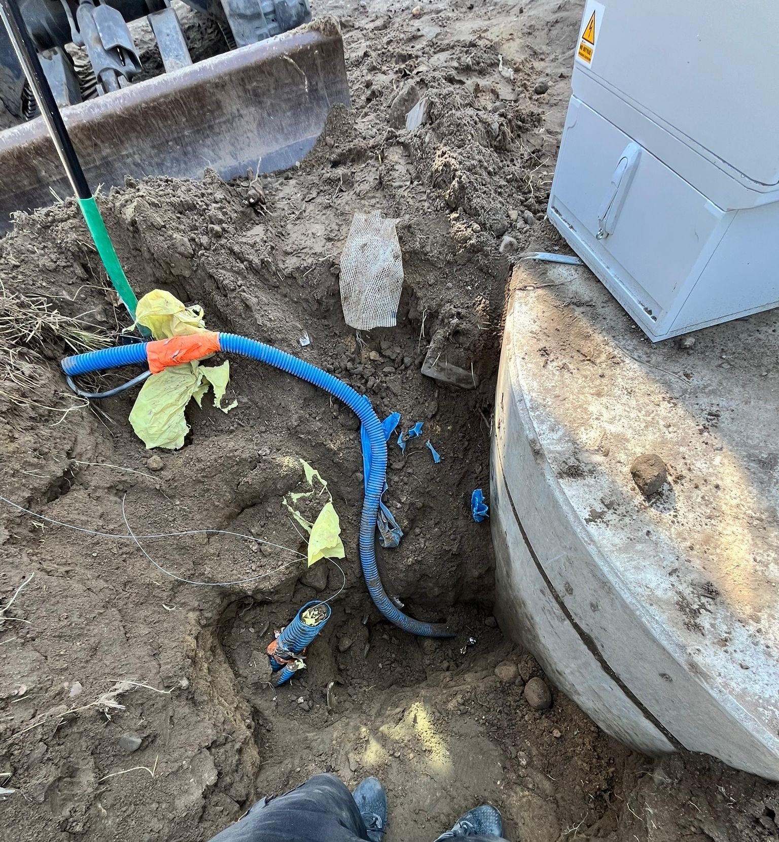 Wykonane przyłącze elektryczne do stacji wodociągowej z widocznymi kablami w ziemi, elementami przepompowni i fragmentem koparki w tle. Prace ziemne w toku.