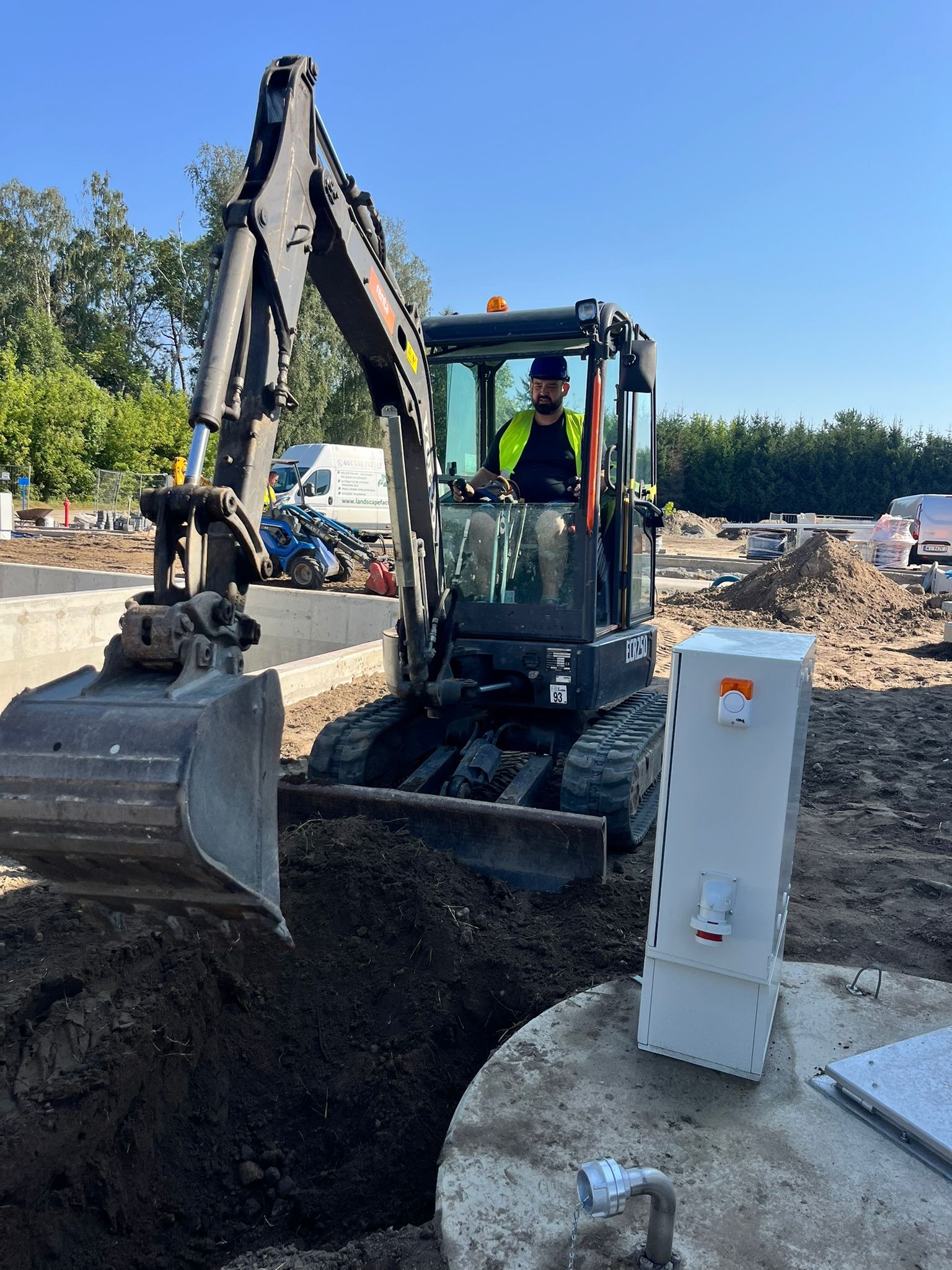 Operator koparki w trakcie prac ziemnych przy instalacji elektrycznej. Widoczna skrzynka z elementami elektrycznymi i wykop w ziemi. Słoneczny dzień.