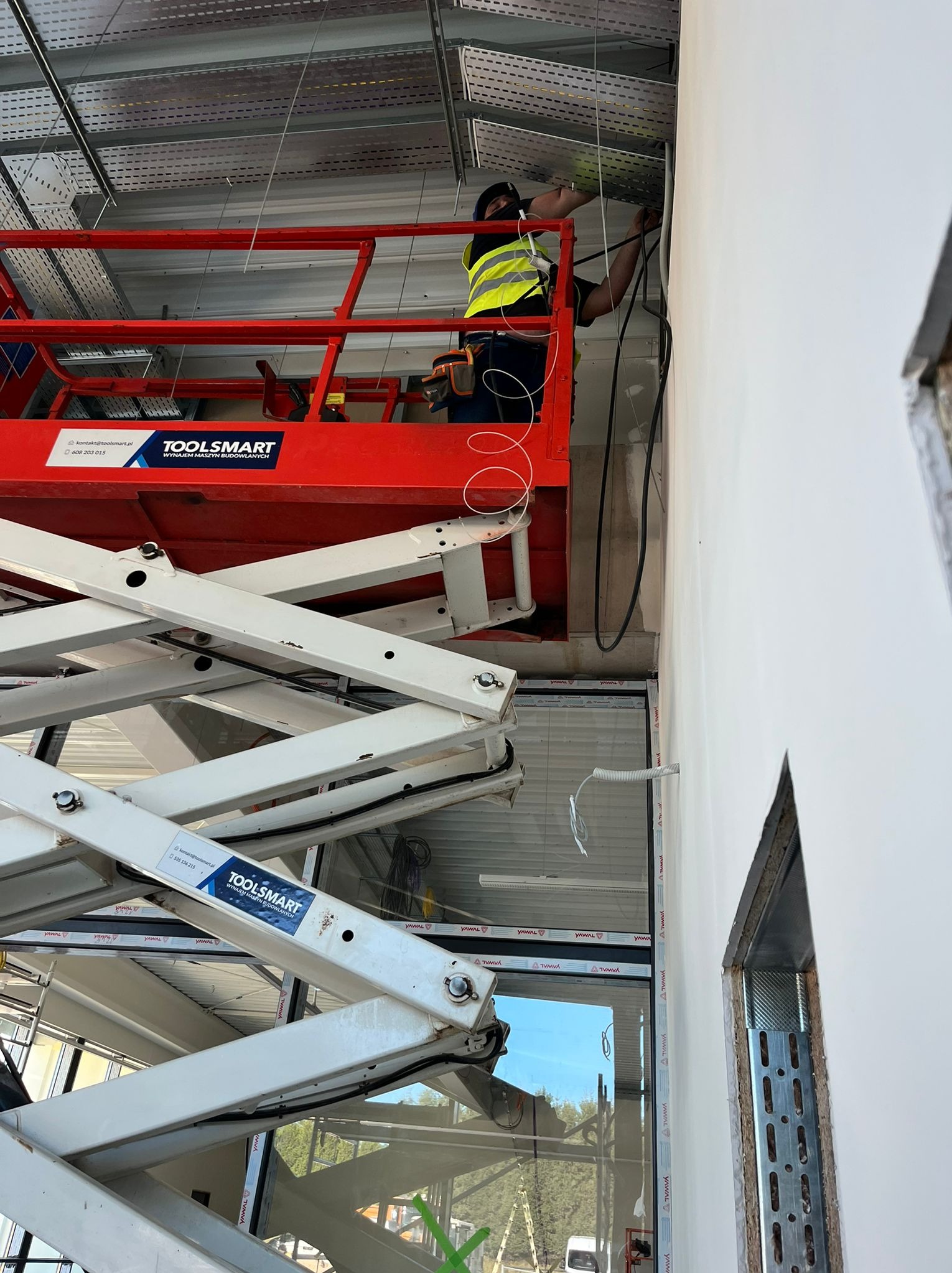 Elektryk w kamizelce na podnośniku montuje kable w metalowej korycie pod sufitem. Widoczne przewody i elementy instalacji. Praca na wysokości.