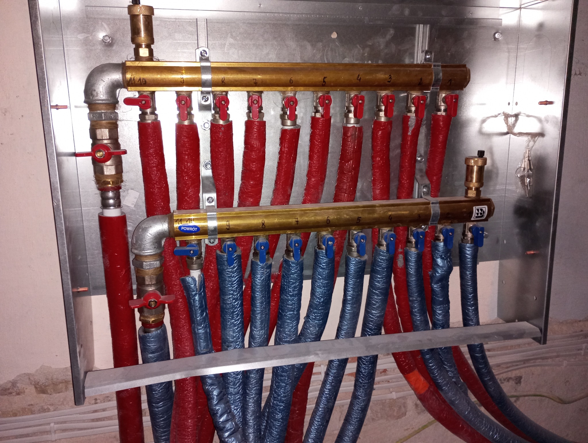 Rozdzielacz ogrzewania podłogowego z rurami w izolacji czerwonej i niebieskiej, z zaworami i numeracją, zamontowany na metalowej płycie. Widoczne przyłącza i fragment ściany.