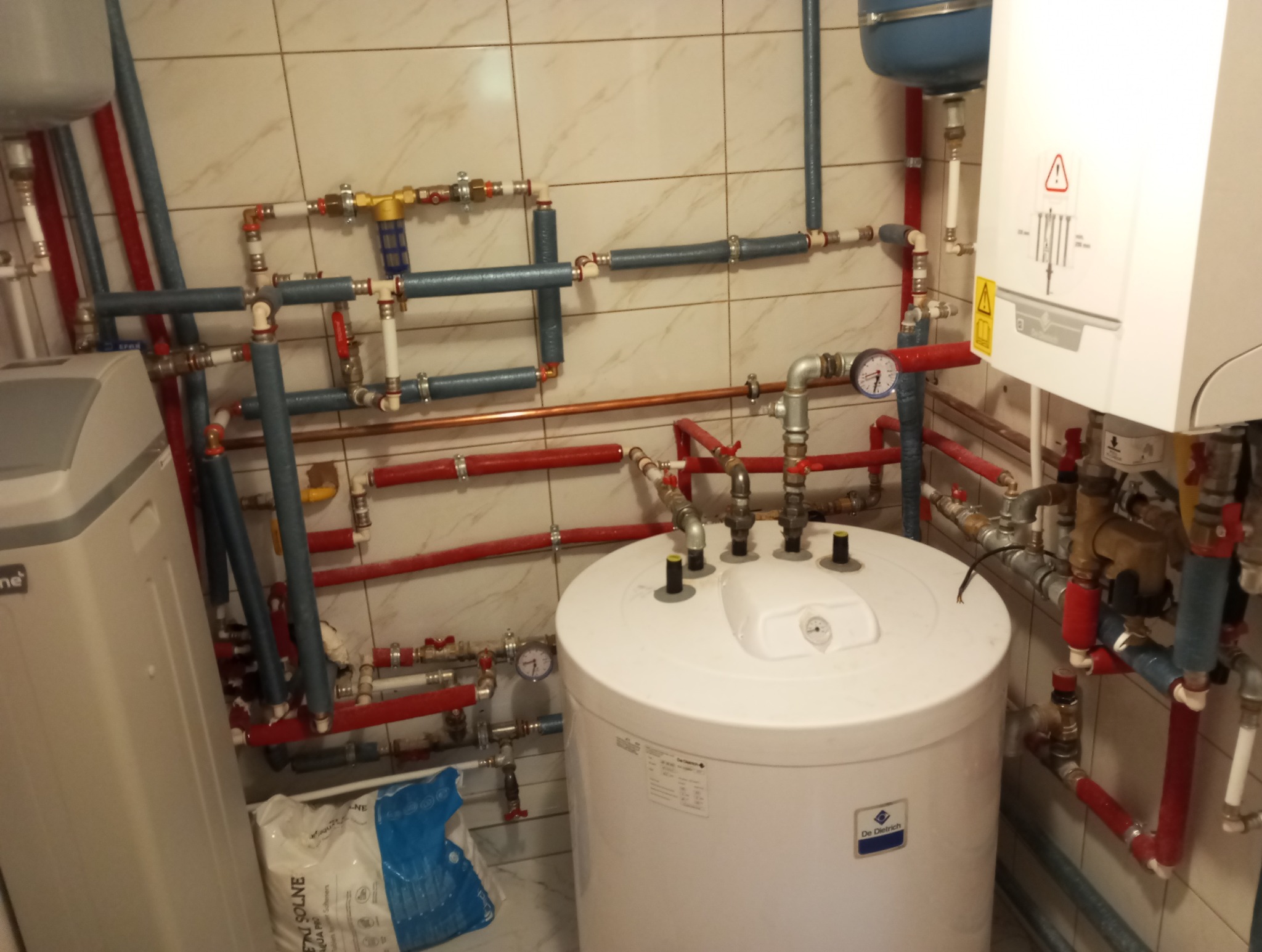 Złożona instalacja CO z piecem gazowym, zbiornikiem i rurami w kolorach czerwonym i niebieskim na tle beżowych płytek w łazience. Widoczne manometry i zawory.