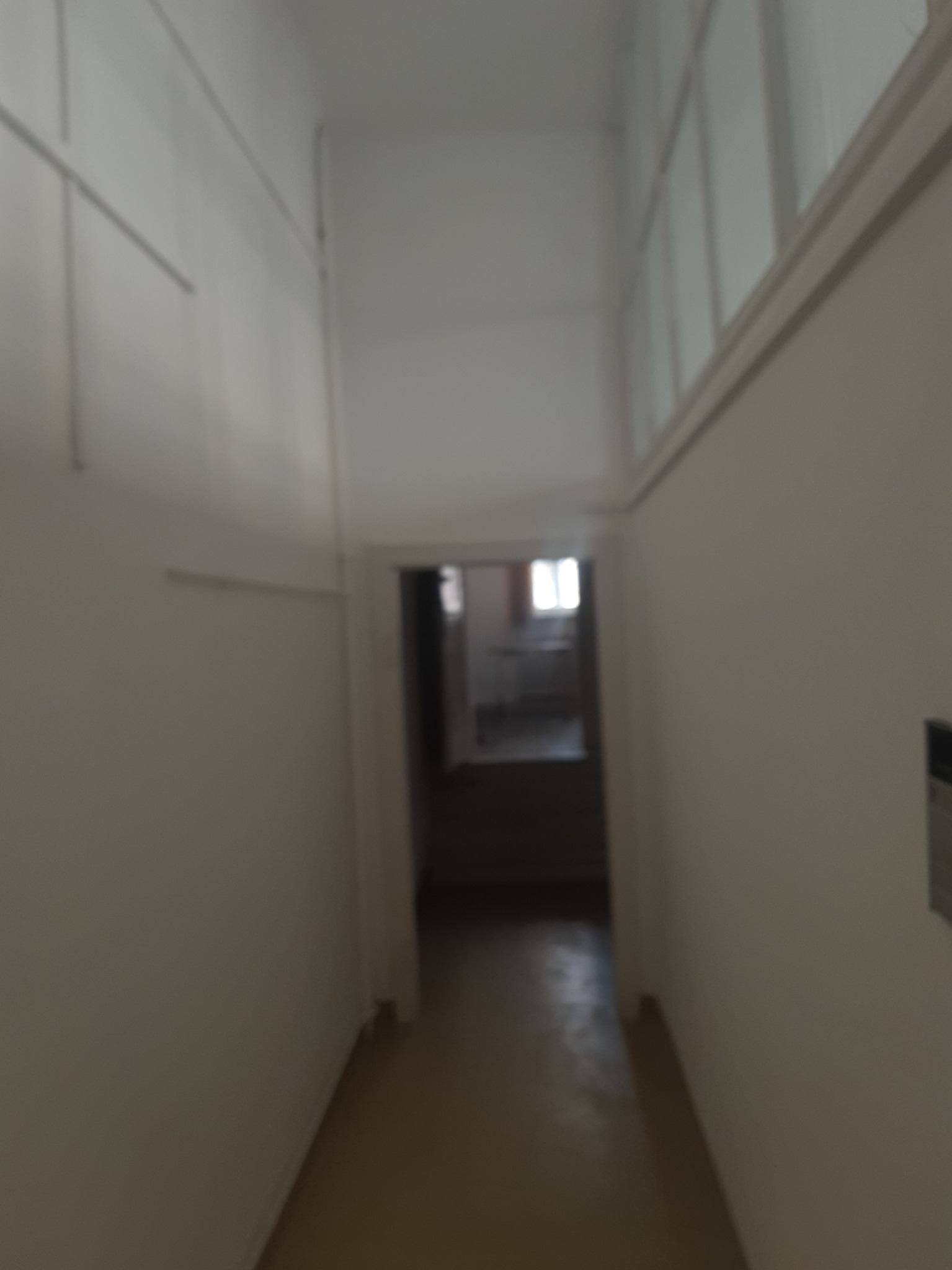 Pomalowany na biało korytarz z widokiem na wnętrze pokoju z oknem. Ściany gładkie, świeżo odmalowane, wpadające światło dzienne.