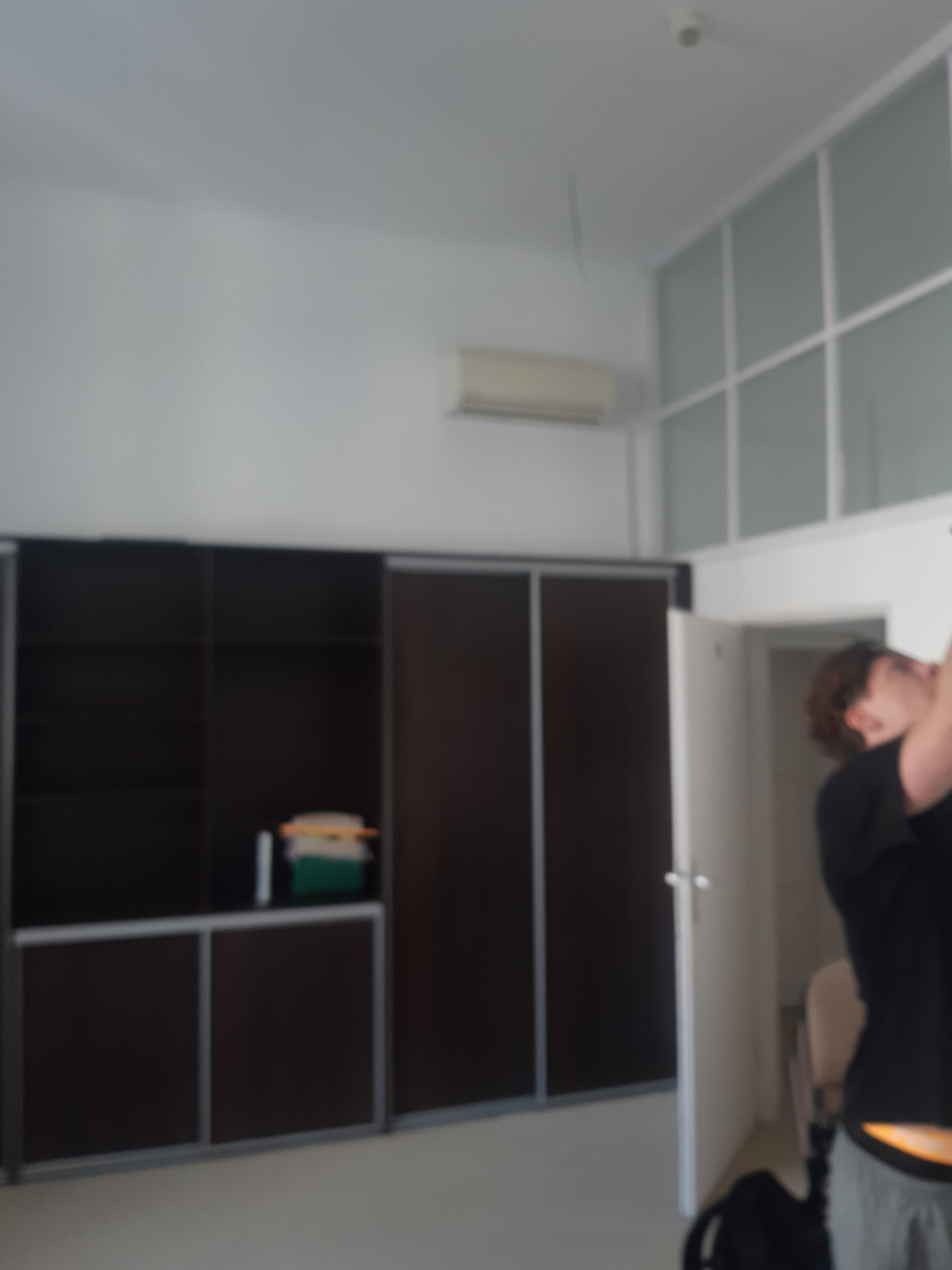 Wnętrze pokoju z dużą, ciemną szafą przesuwną, klimatyzatorem i oknem z białymi ramami. Osoba w tle, prawdopodobnie podczas prac wykończeniowych.