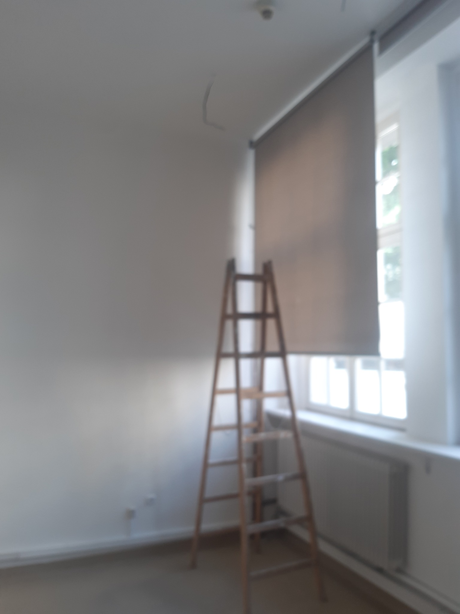Wnętrze pokoju przed pracami wykończeniowymi. Drewniana drabina oparta o ścianę, okno z roletą. Widoczne ślady po instalacji elektrycznej na ścianie.