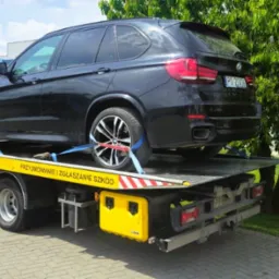 Czarny SUV BMW na żółtej lawecie pomocy drogowej, zabezpieczony pasami, z widocznym numerem rejestracyjnym na tle zieleni.